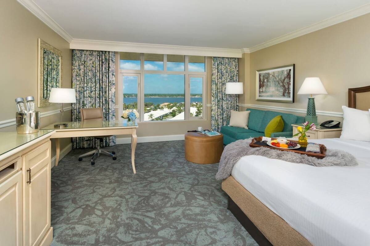 A hotel room with a desk, a bed, a sofa, a view of the ocean, and a painting on the wall.