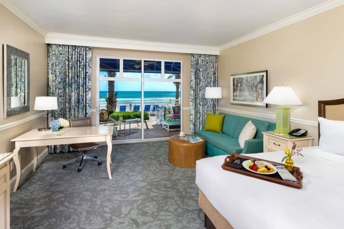 A hotel room with a bed, a desk, a chair, a sofa, a small table, a nightstand, a lamp, a tray with food, and a view of the ocean.