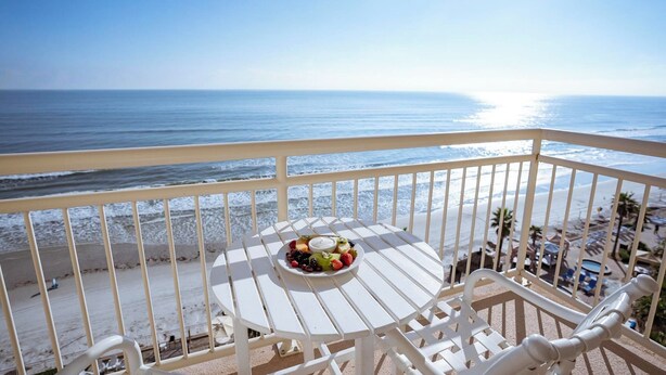 Un balcón con una mesa redonda y una bandeja de frutas, con vista a una playa y cielos despejados.