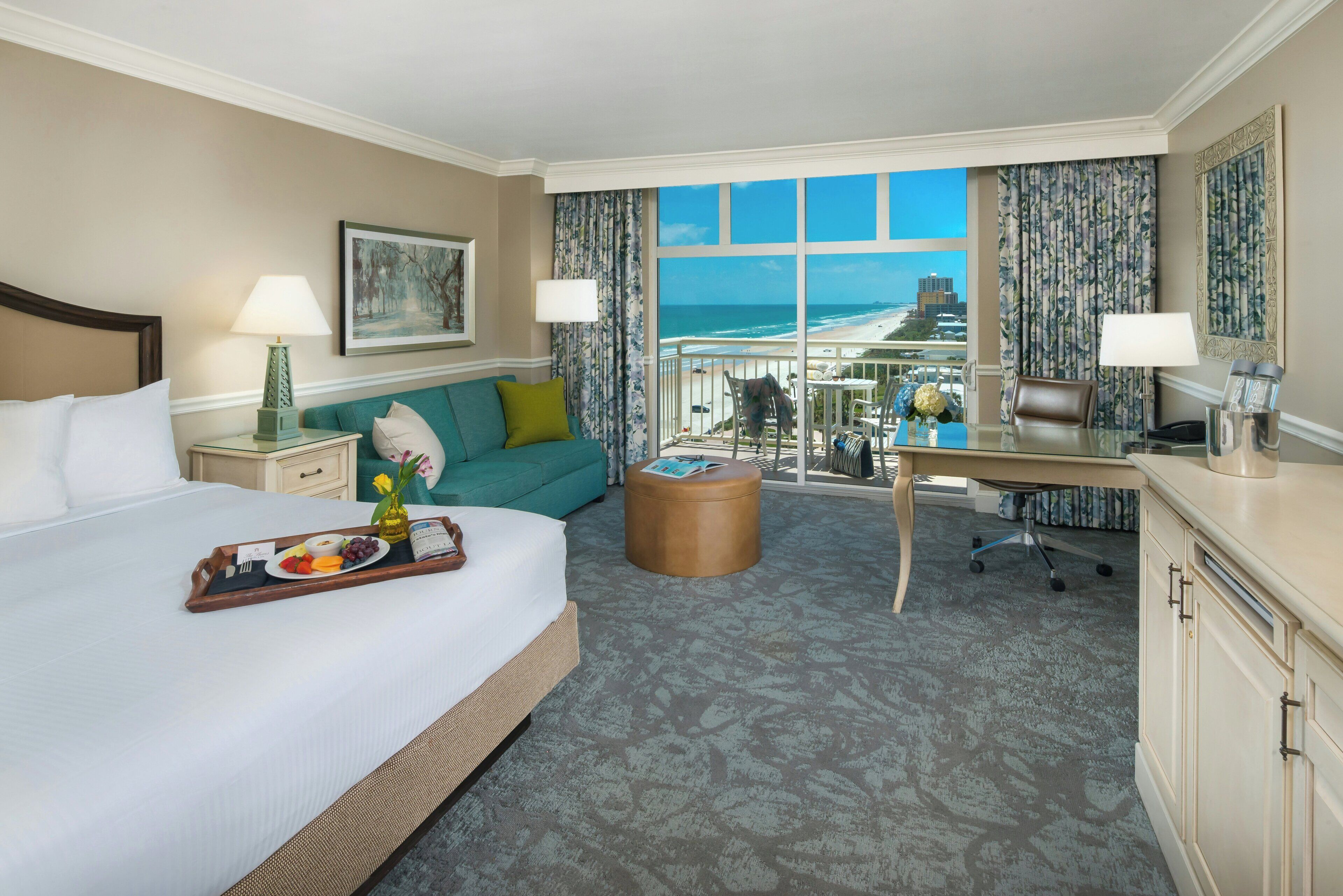 A hotel room with a bed, a desk, a balcony with a view of the beach, and a dining trolley with fruit and a bottle.