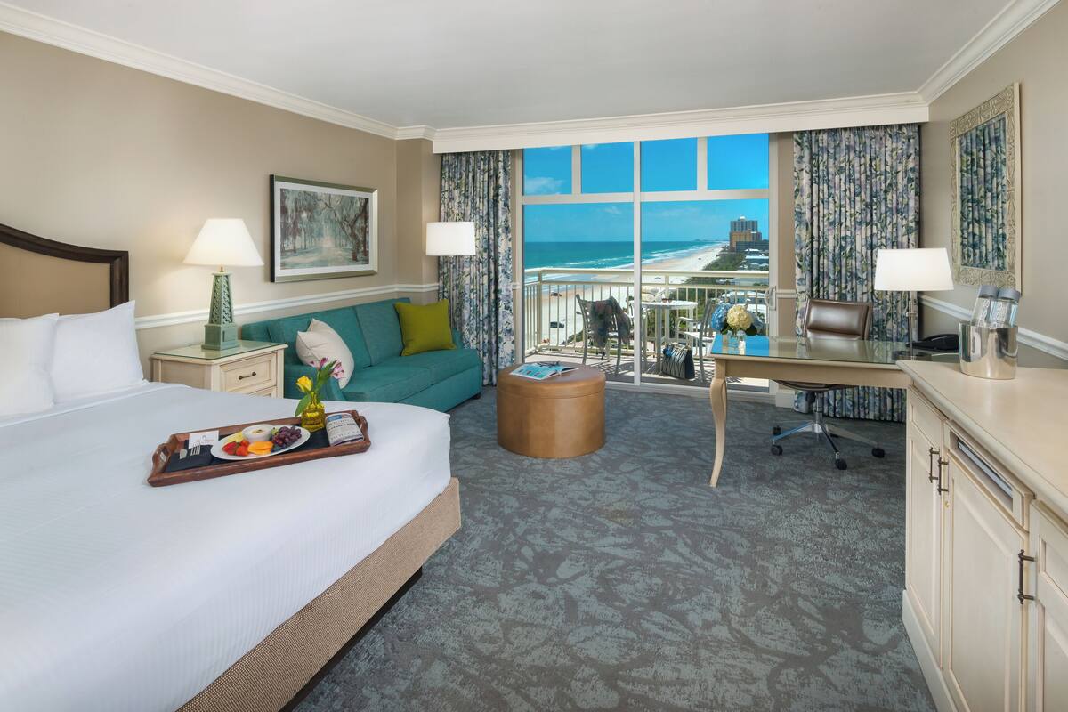 A hotel room with a bed, a desk, a balcony with a view of the beach, and a dining table with a tray of food.