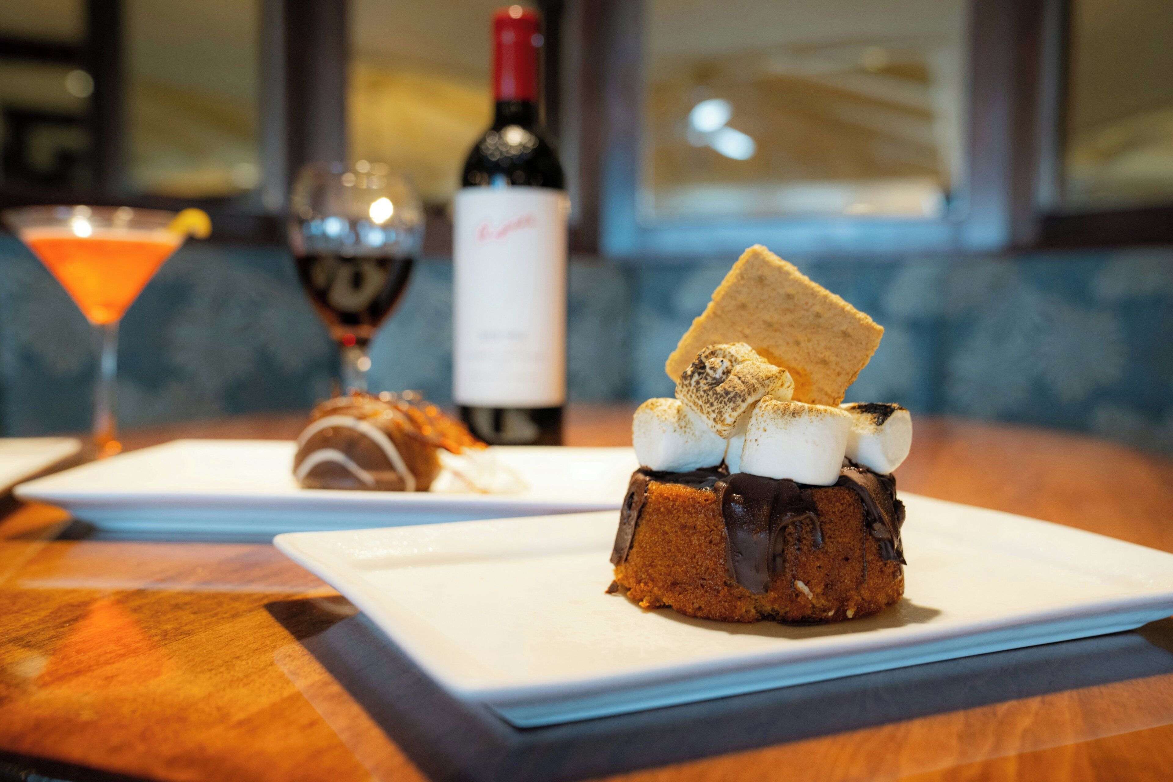 A dessert with marshmallows and chocolate on a plate, a bottle of wine, and a martini glass on a table.