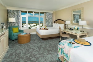 Habitación de hotel con cama, escritorio, silla, reposapiés pequeño y vista al mar.