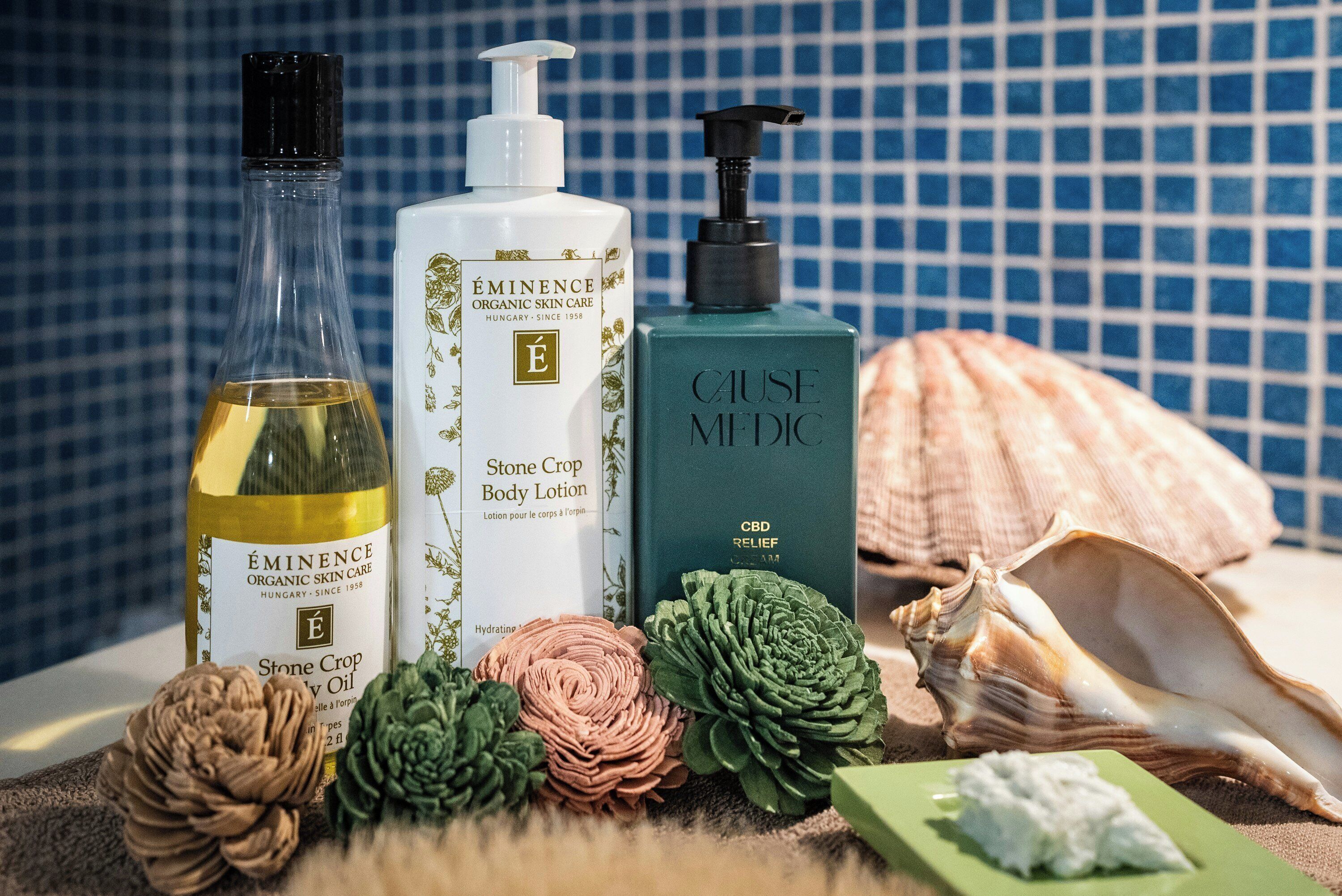 Una selección de productos para el cuidado de la piel, incluyendo una botella de aceite, loción corporal y crema, junto con elementos decorativos como conchas marinas y una planta verde.