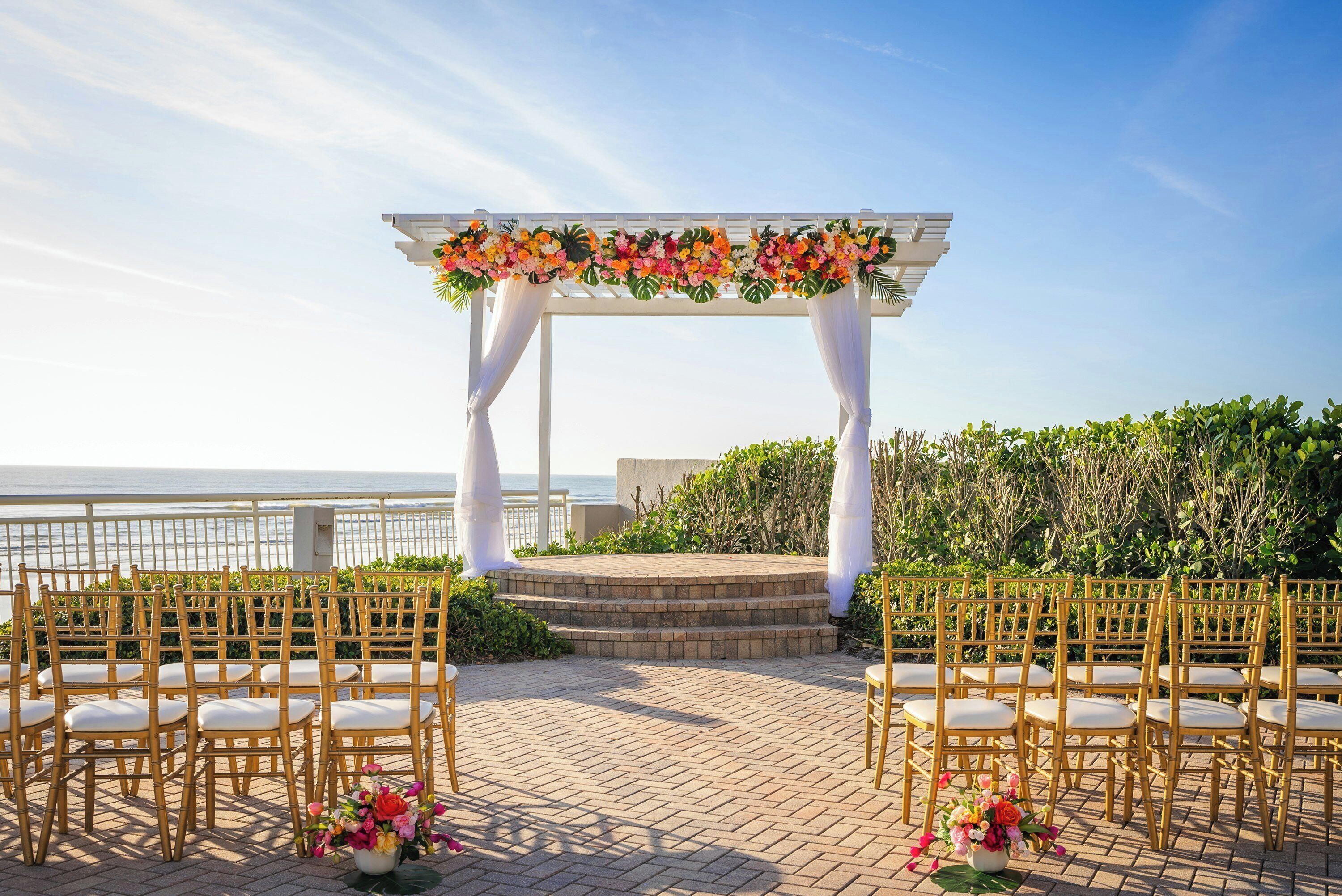Cerimônia de casamento ao ar livre com um arco branco e decorações florais, com vista para um corpo de água.