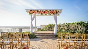 Cerimônia de casamento ao ar livre com um arco branco e decorações florais, com vista para um corpo de água.