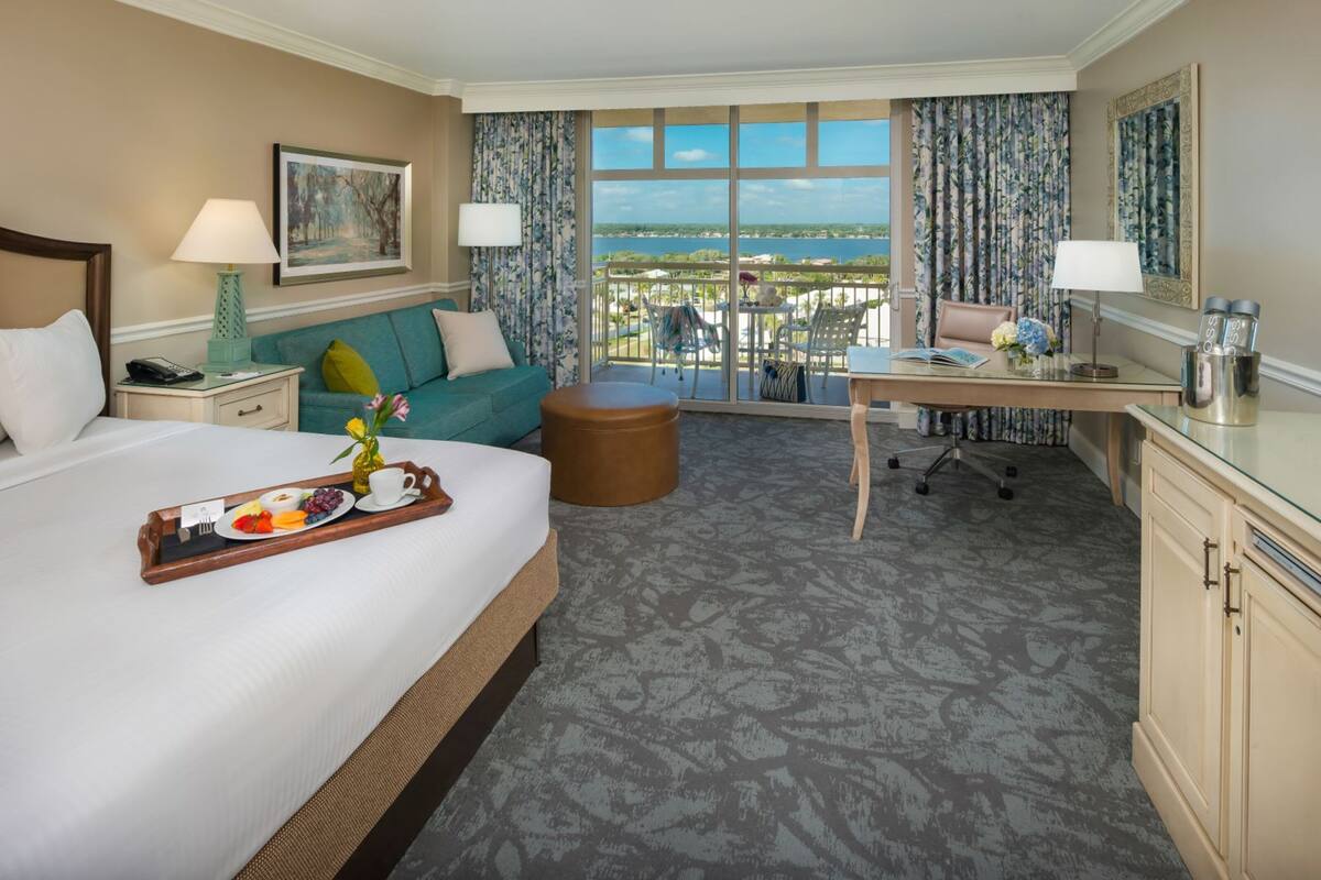 A hotel room with a large bed, a desk, a balcony with a view, and a dining table with breakfast.