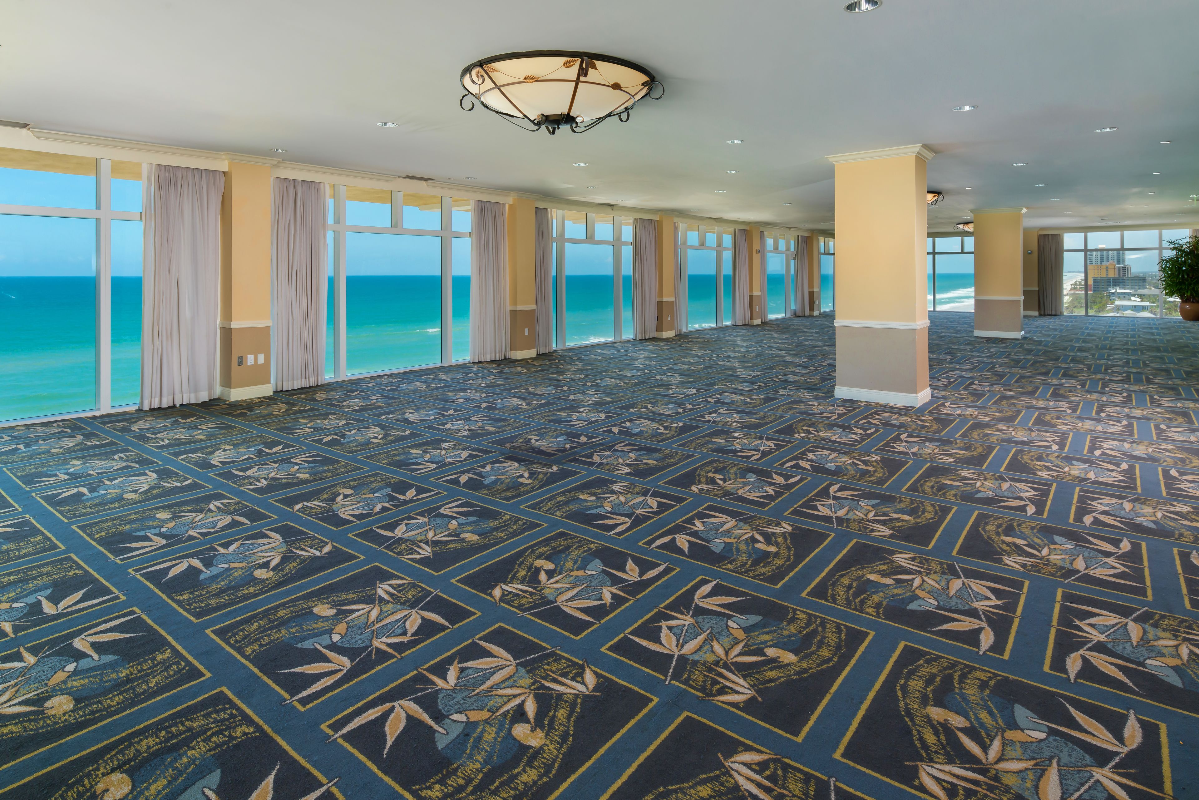 A spacious hotel lobby with a patterned carpet, large windows with curtains, and a view of the ocean.