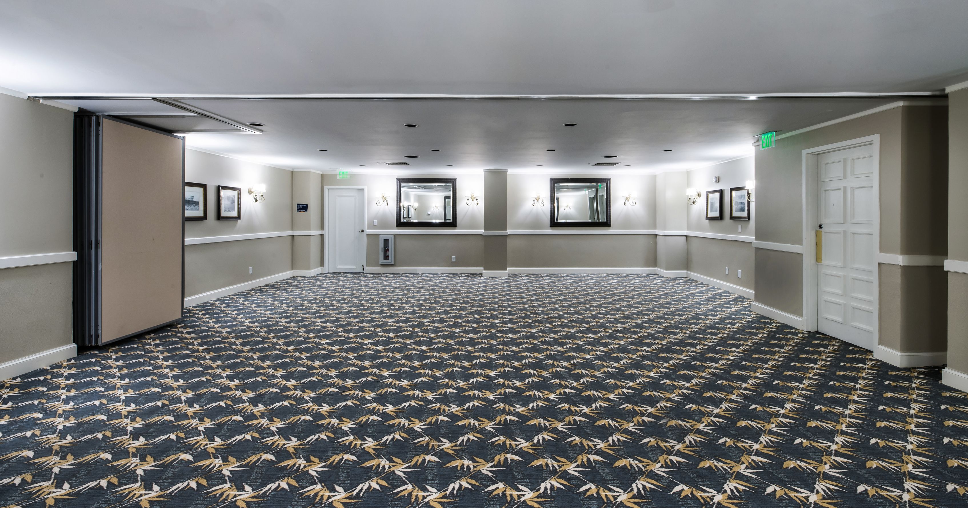 A hotel hallway with a patterned carpet, framed pictures on the walls, and a door on the right.