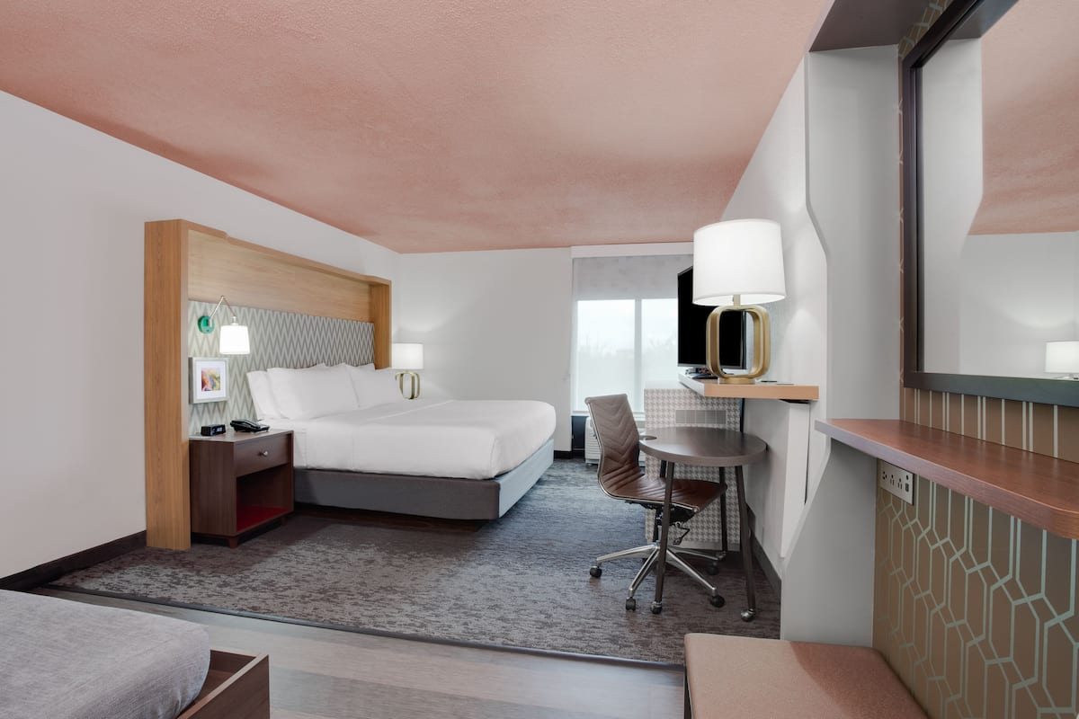 A hotel room with a bed, bedside table, lamp, desk, chair, and a large mirror.
