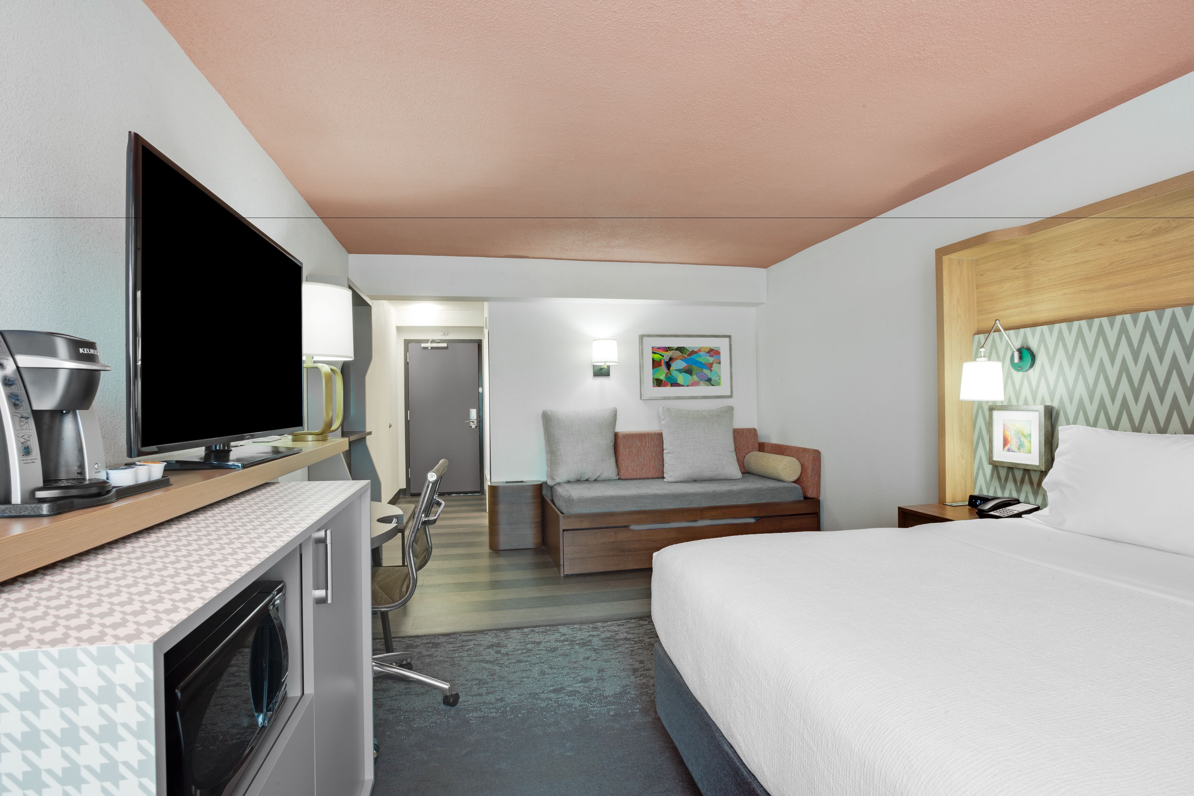 A hotel room with a bed, a TV, a desk with a coffee maker, a chair, and a small kitchenette.