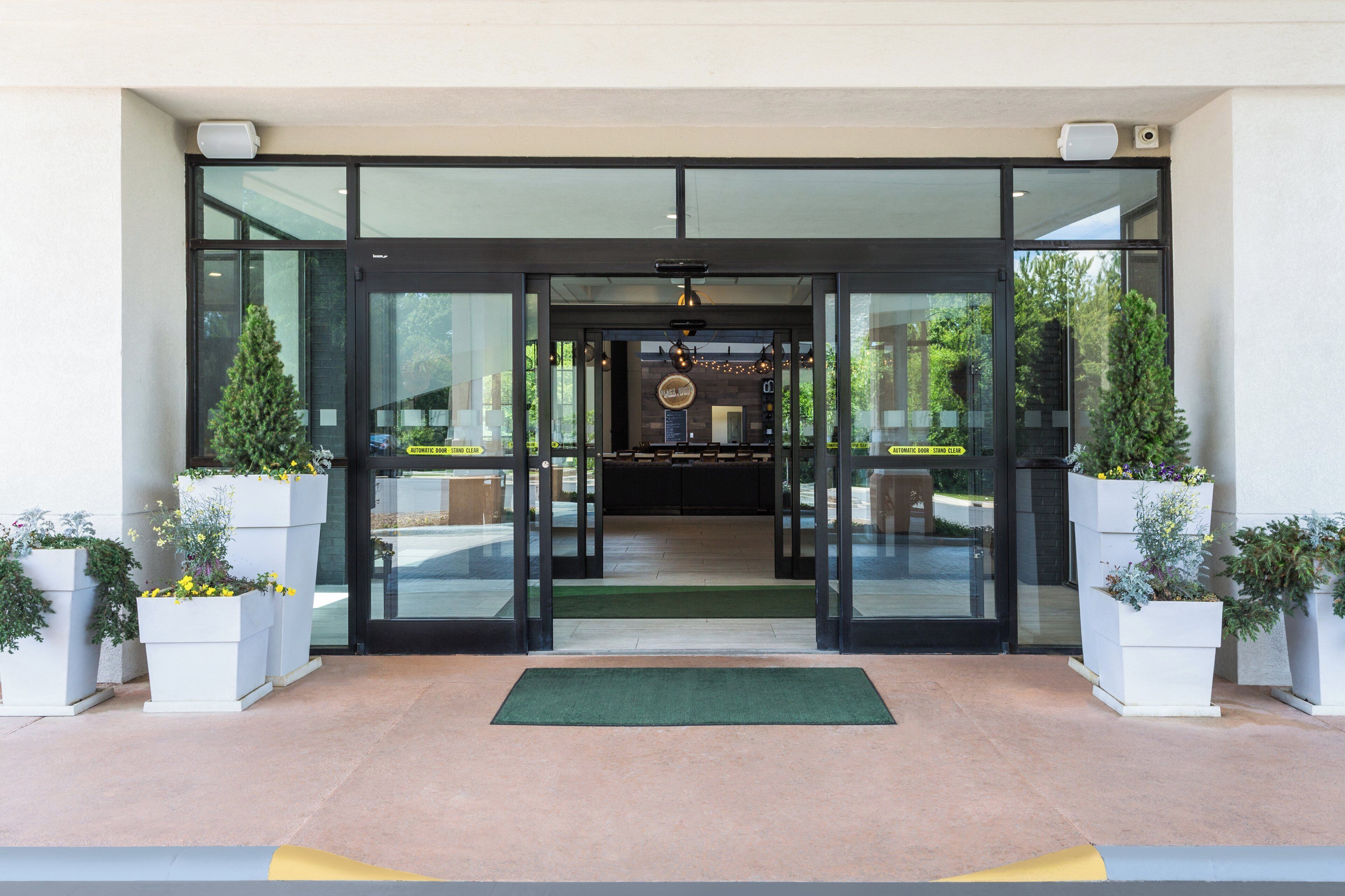 A hotel entrance with automatic doors, a green mat, and potted plants.