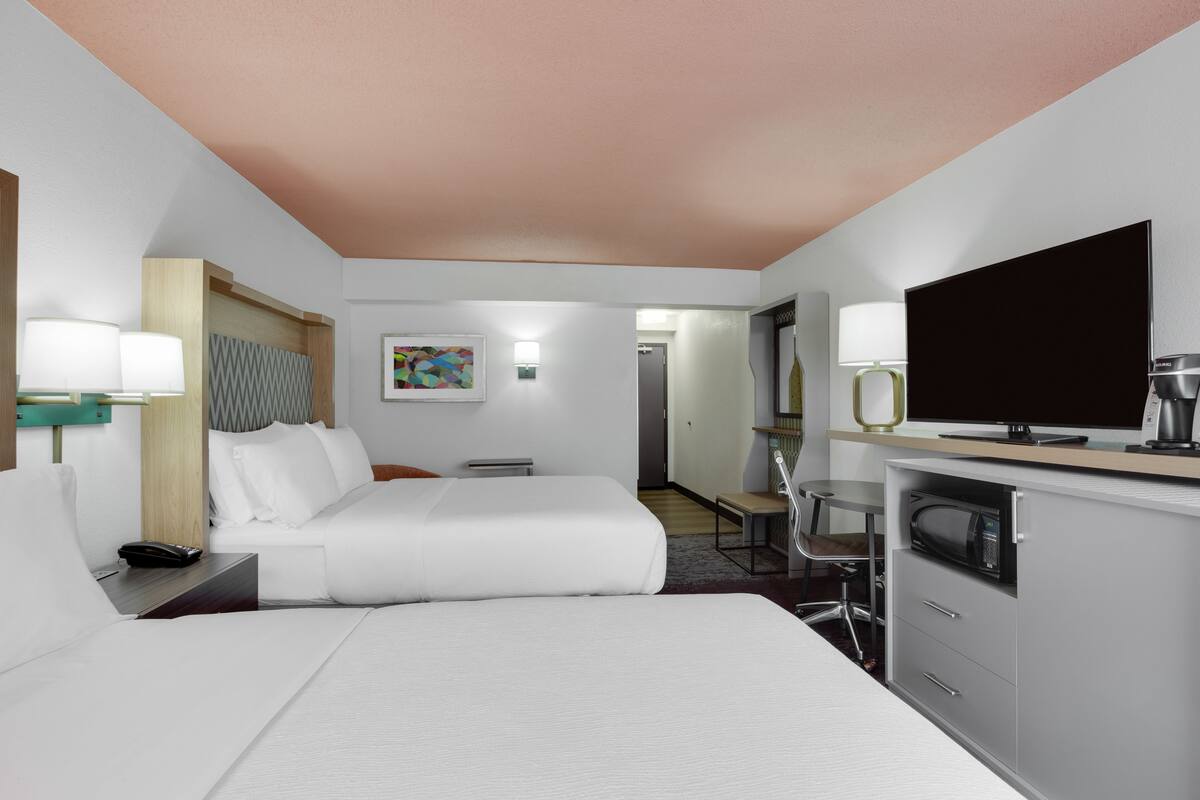 A hotel room with a bed, bedside table, lamp, TV, microwave, and a small desk area.