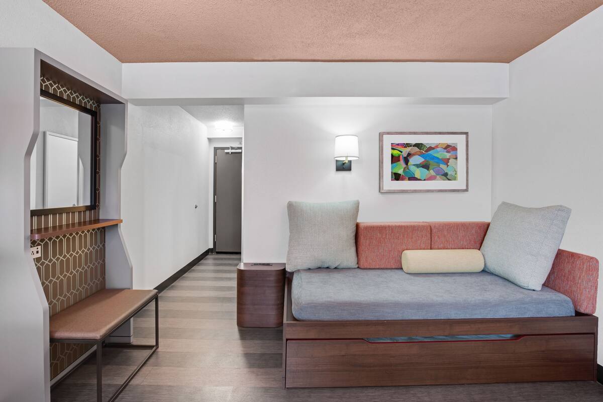 A modern hotel room with a wooden bench, a sofa with cushions, a wall-mounted light, and a colorful abstract painting.