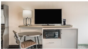 Ein kleines Hotelzimmer mit Schreibtisch, Stuhl, Fernseher, Lampe und einer Kaffeemaschine.