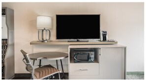 Une chambre d’hôtel de taille modeste, équipée d’un bureau, d’une chaise, d’une télévision, d’une lampe et d’une machine à café.