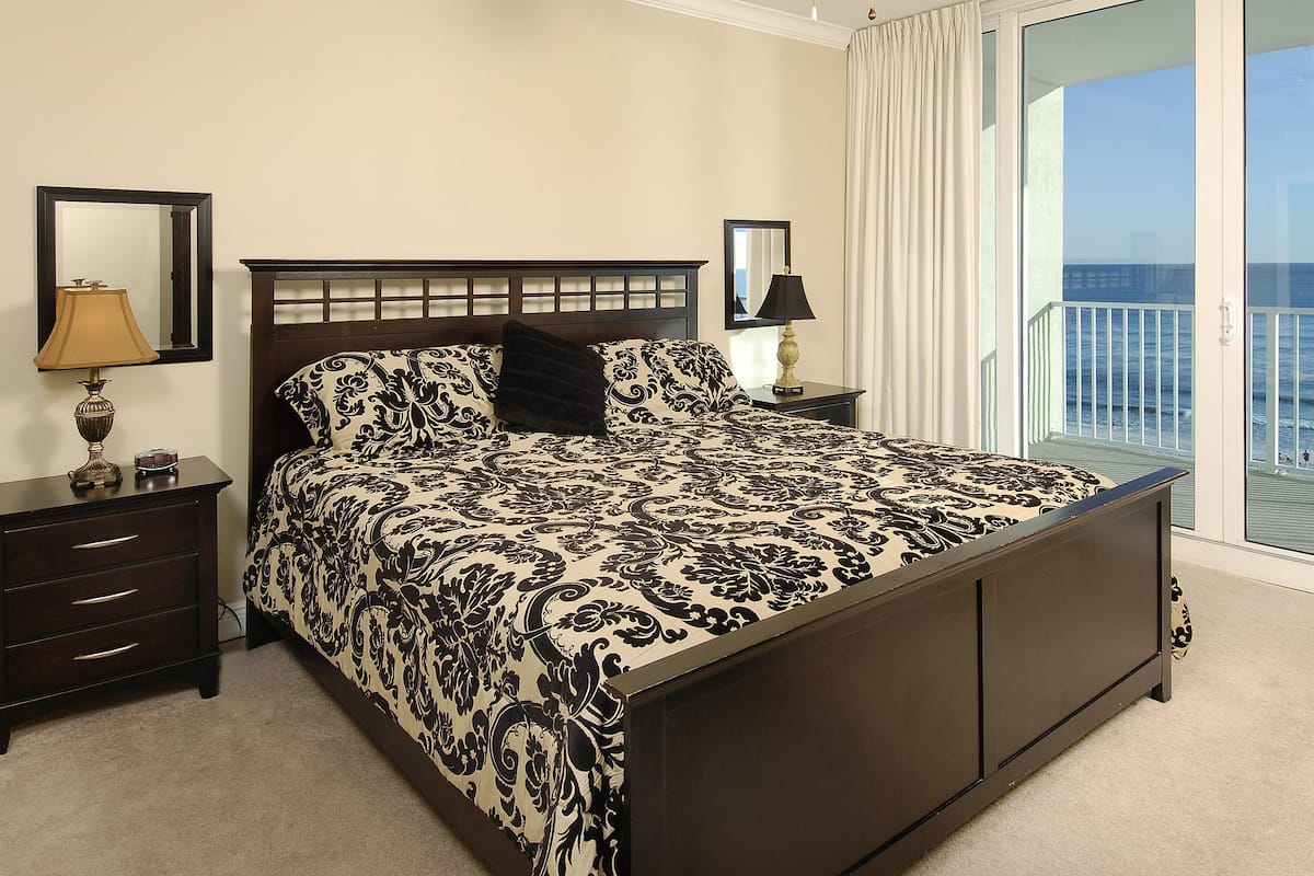 A bedroom with a large bed, a nightstand with a lamp, a dresser, and a balcony with a view of the ocean.
