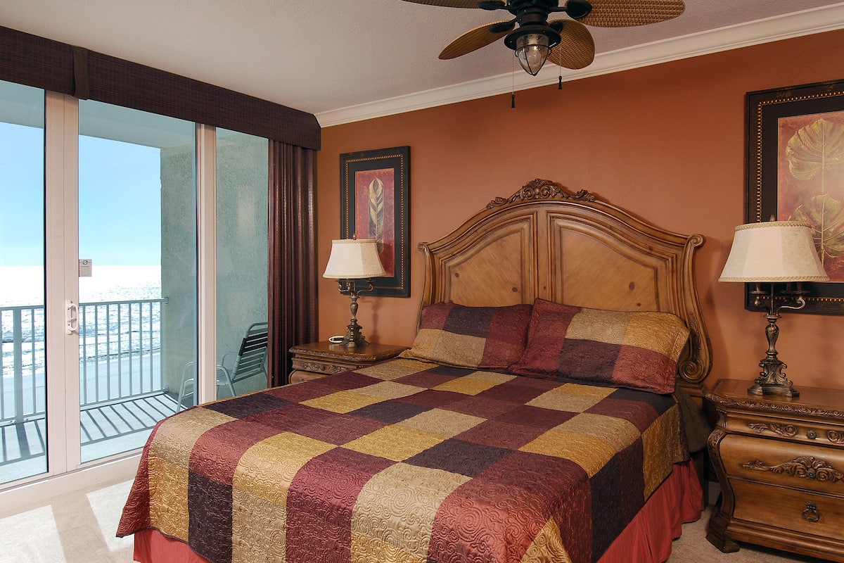 A hotel room with a large bed, two bedside tables, a ceiling fan, and a view of the beach through the sliding glass door.