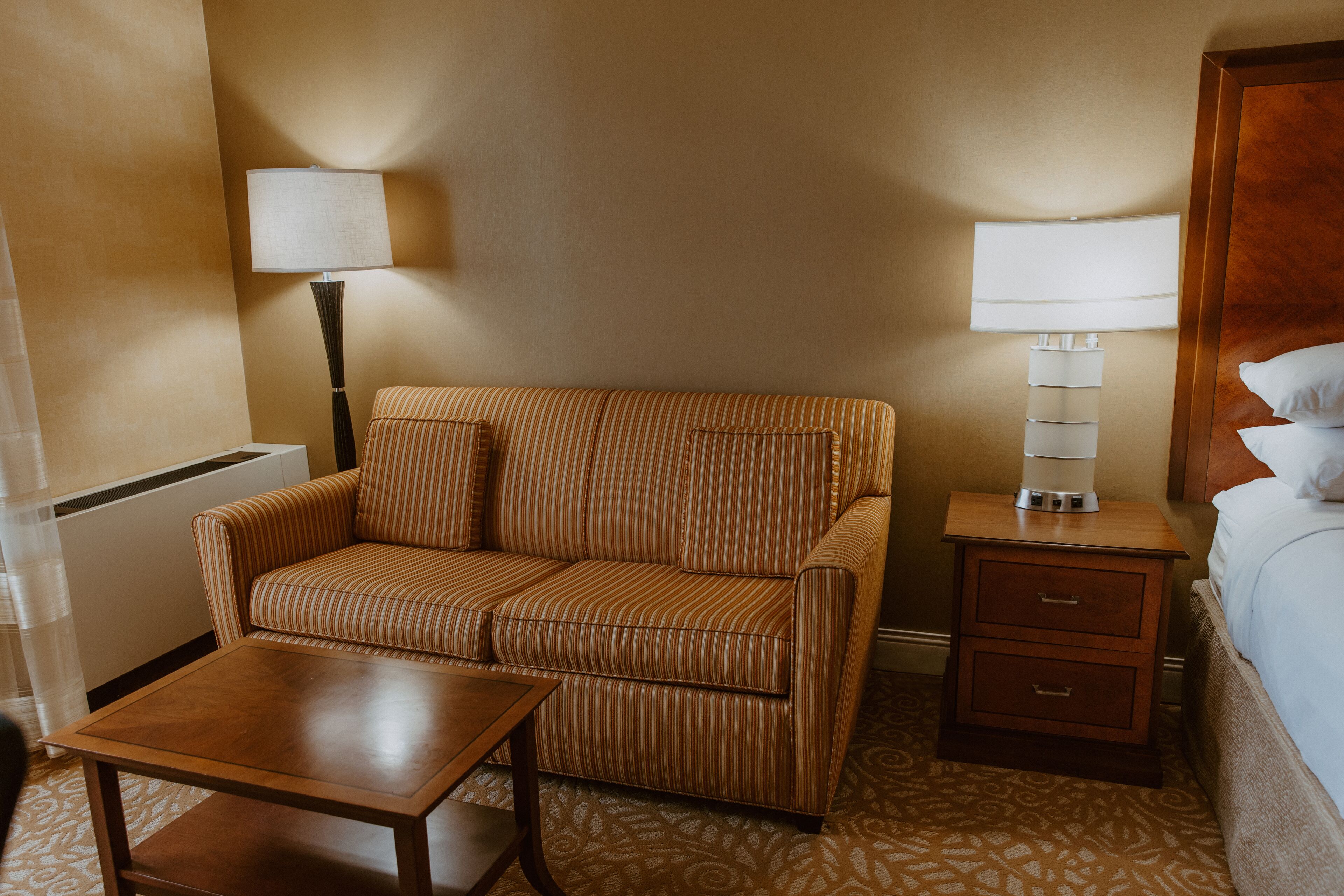 A hotel room with a striped sofa, a wooden nightstand with a lamp, a small table, and a bed with white linens.