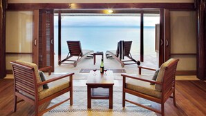 A room with a wooden table, four chairs, and a bottle of wine. The room has a view of the ocean through large glass doors.