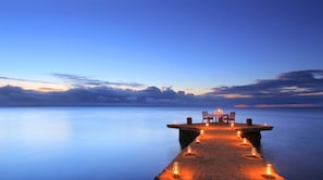 A pier extending into calm waters with a table and chairs set for dining, surrounded by candles.