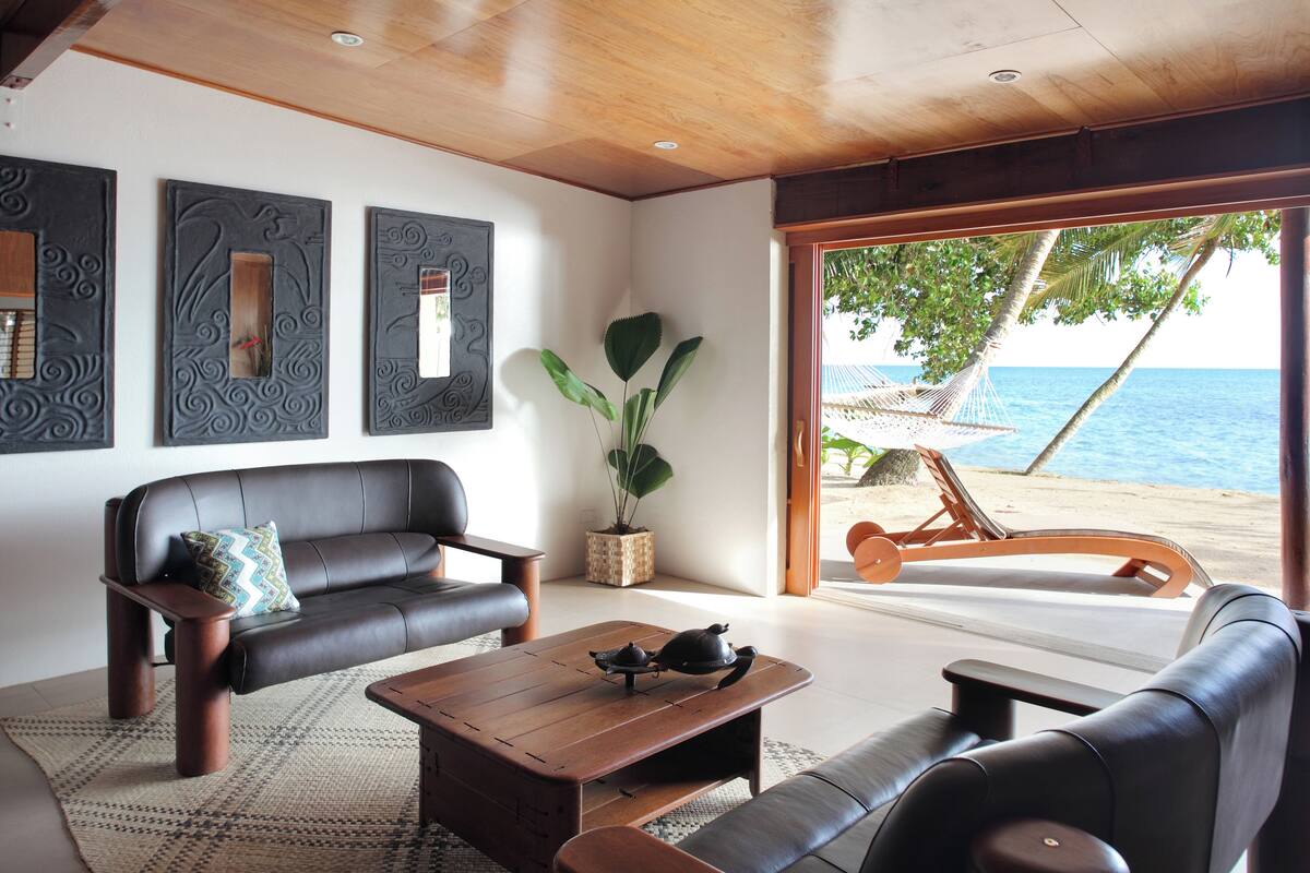 Un salon moderne avec un canapé, une table basse et des œuvres d’art décoratives au mur. Une grande fenêtre offre une vue sur l’océan et sur des chaises de plage.