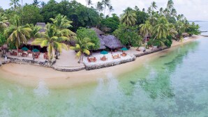 A tropical beach resort with thatched-roof huts, palm trees, and outdoor seating by the water.