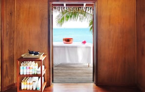 A wooden room with a view of the sea, a table with a bowl and a flower, and shelves with bottles.