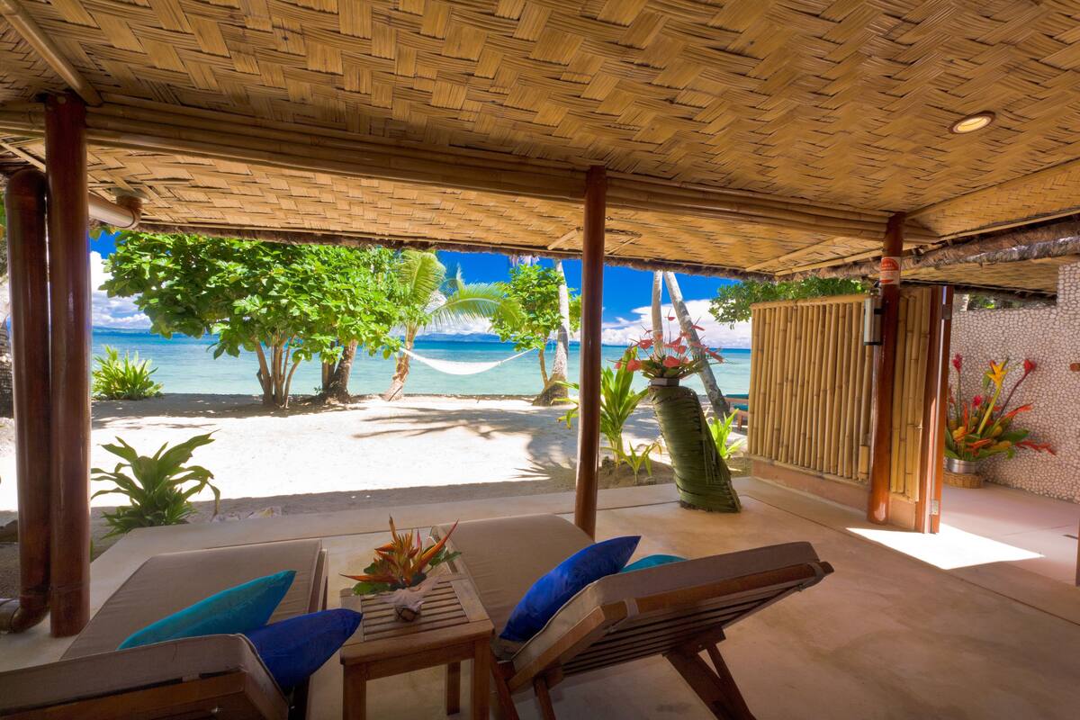 Un salon de plage avec vue sur l’océan, comprenant des chaises longues, une table et des plantes tropicales.
