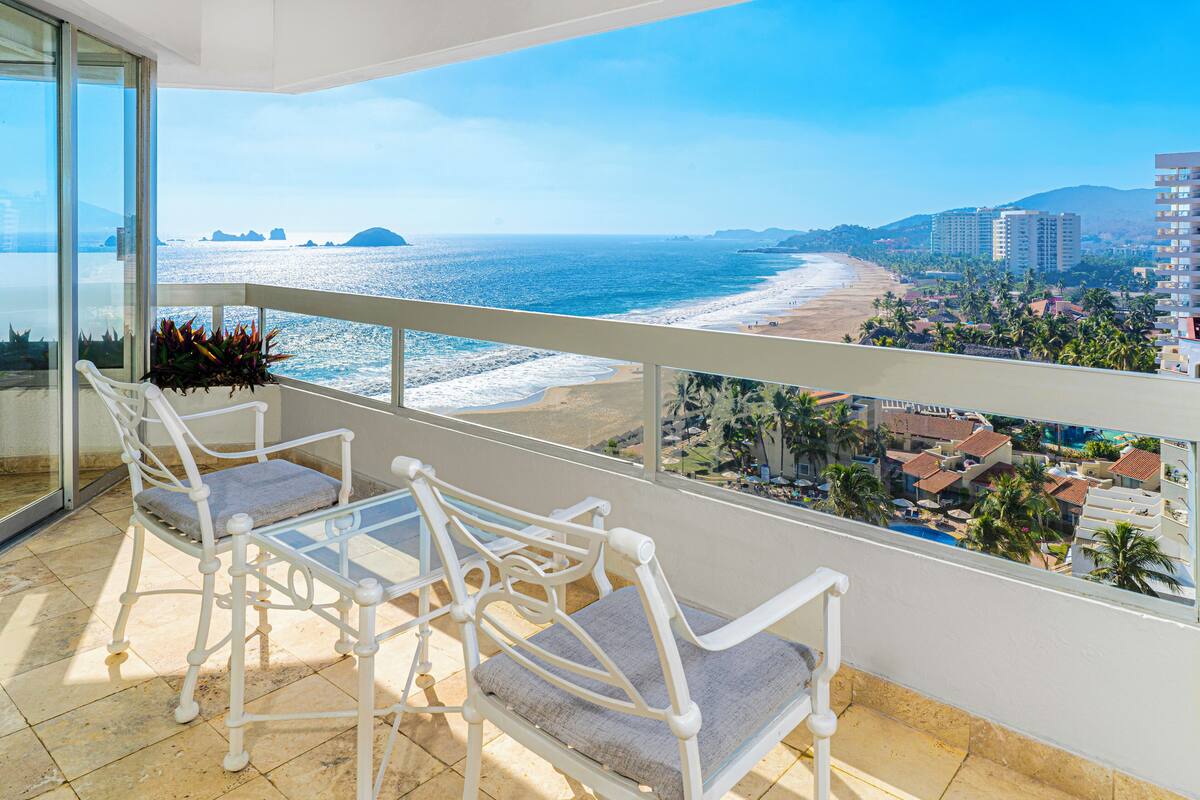 Un balcon avec des chaises en métal blanc et une table en verre, offrant une vue sur une plage et le paysage urbain.