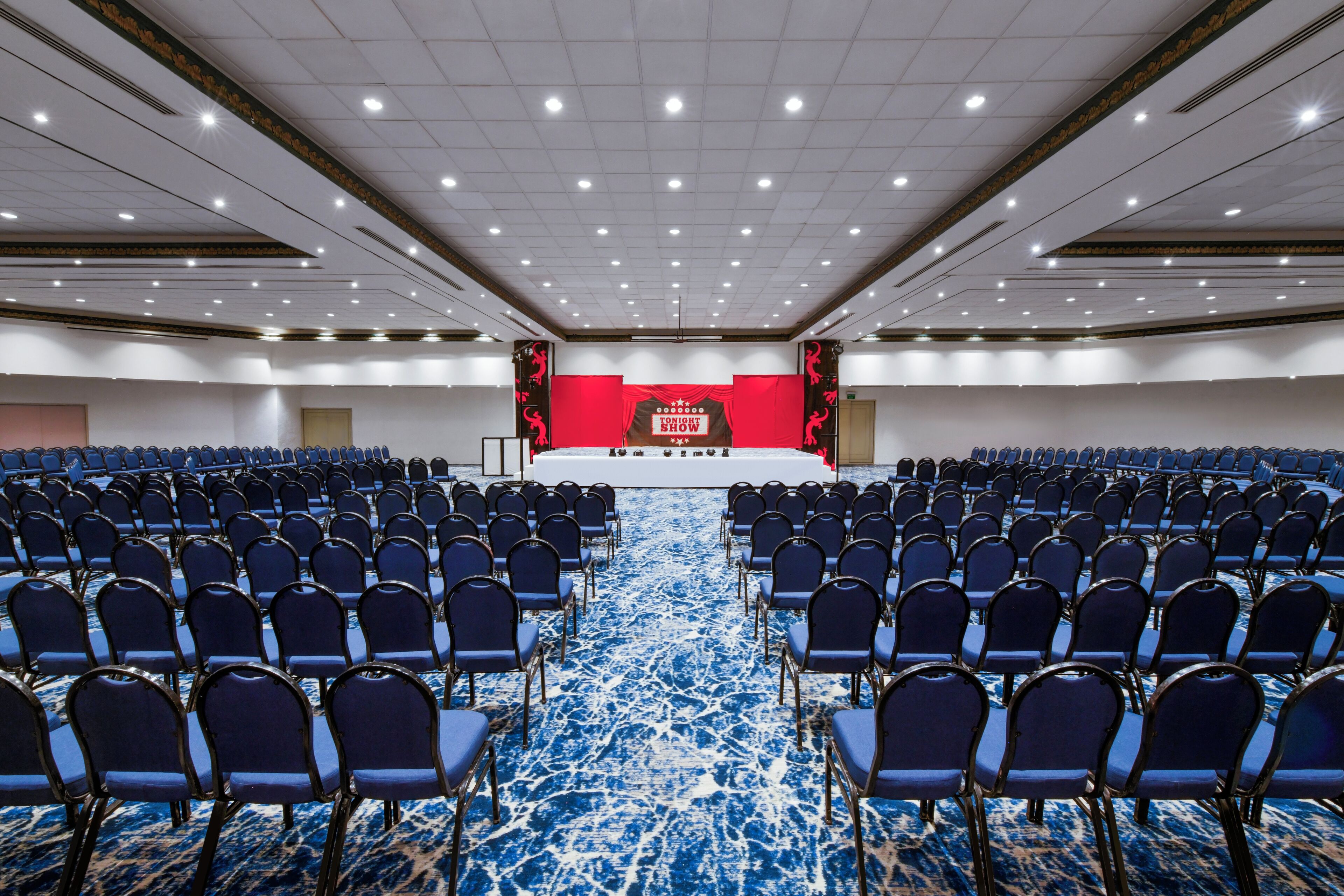 A large conference room with rows of blue chairs facing a stage with a red backdrop.