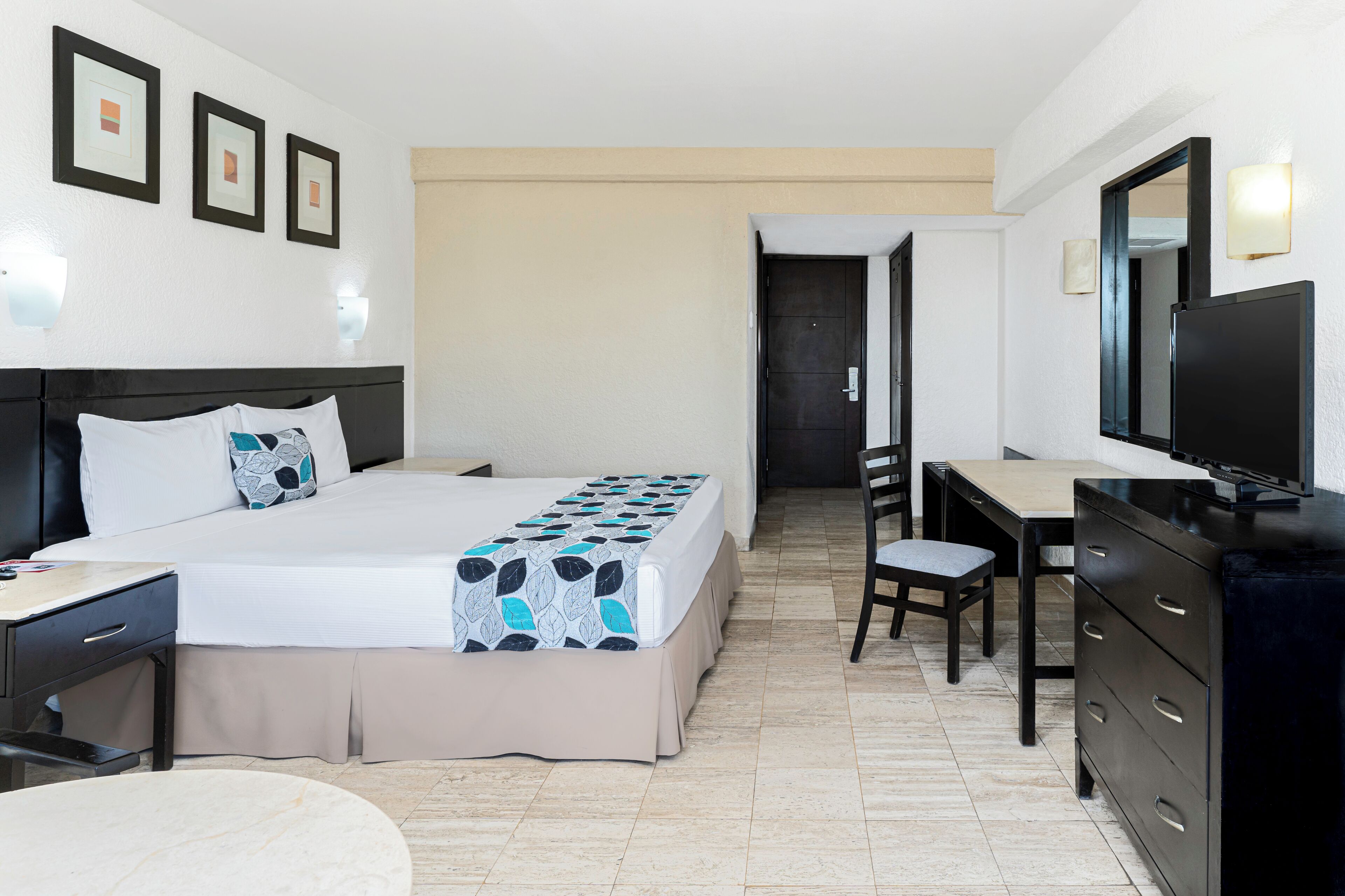 A hotel room with a bed, desk, chair, dresser, and a TV.