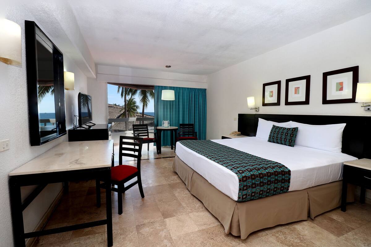 Une chambre d’hôtel avec un lit, un bureau, des chaises, une petite table et une vue sur l’océan.