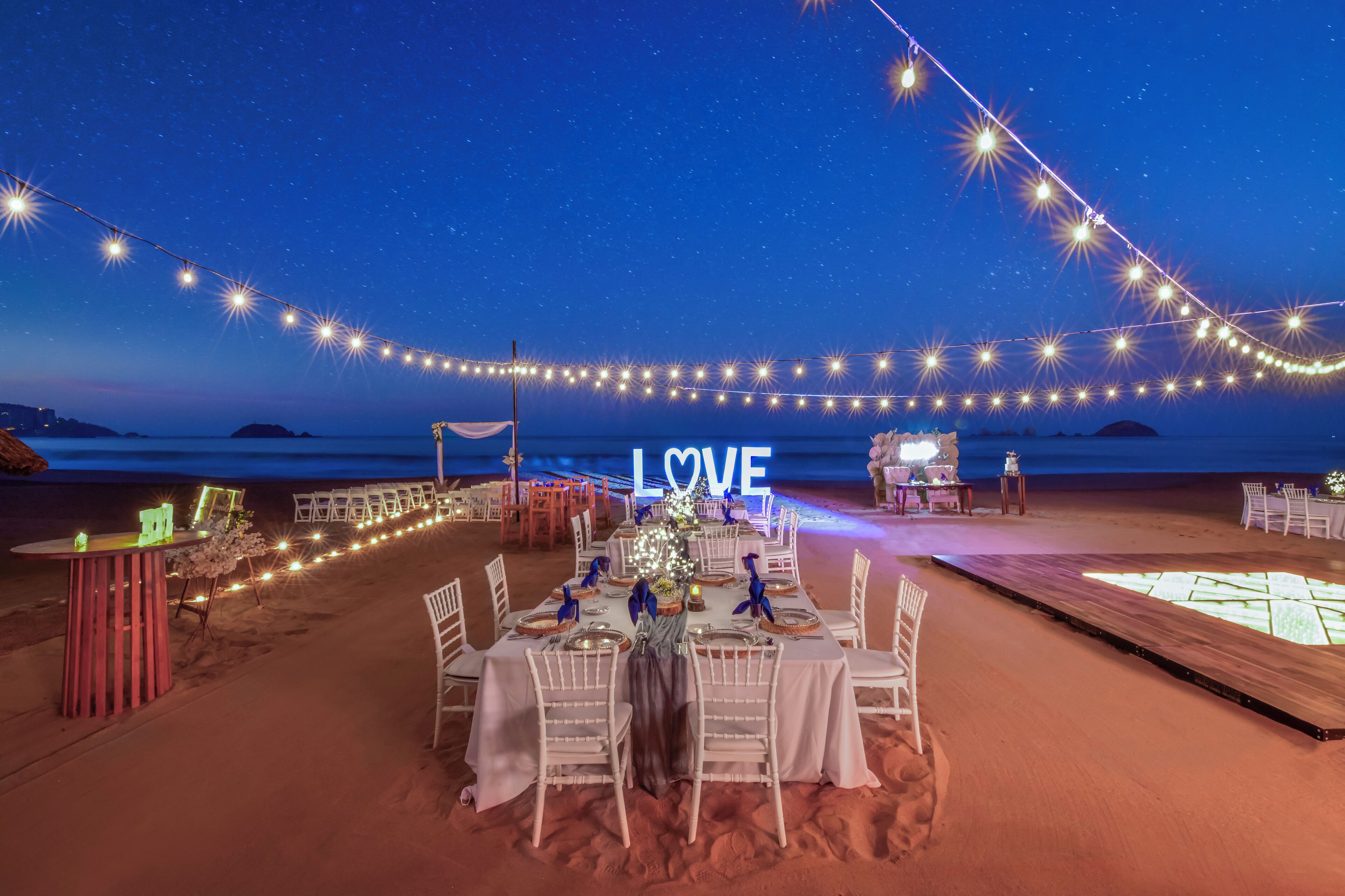 Un'area ristorazione in spiaggia con luci a stringa, un cartello con la scritta 'LOVE' e pavimento sabbioso.