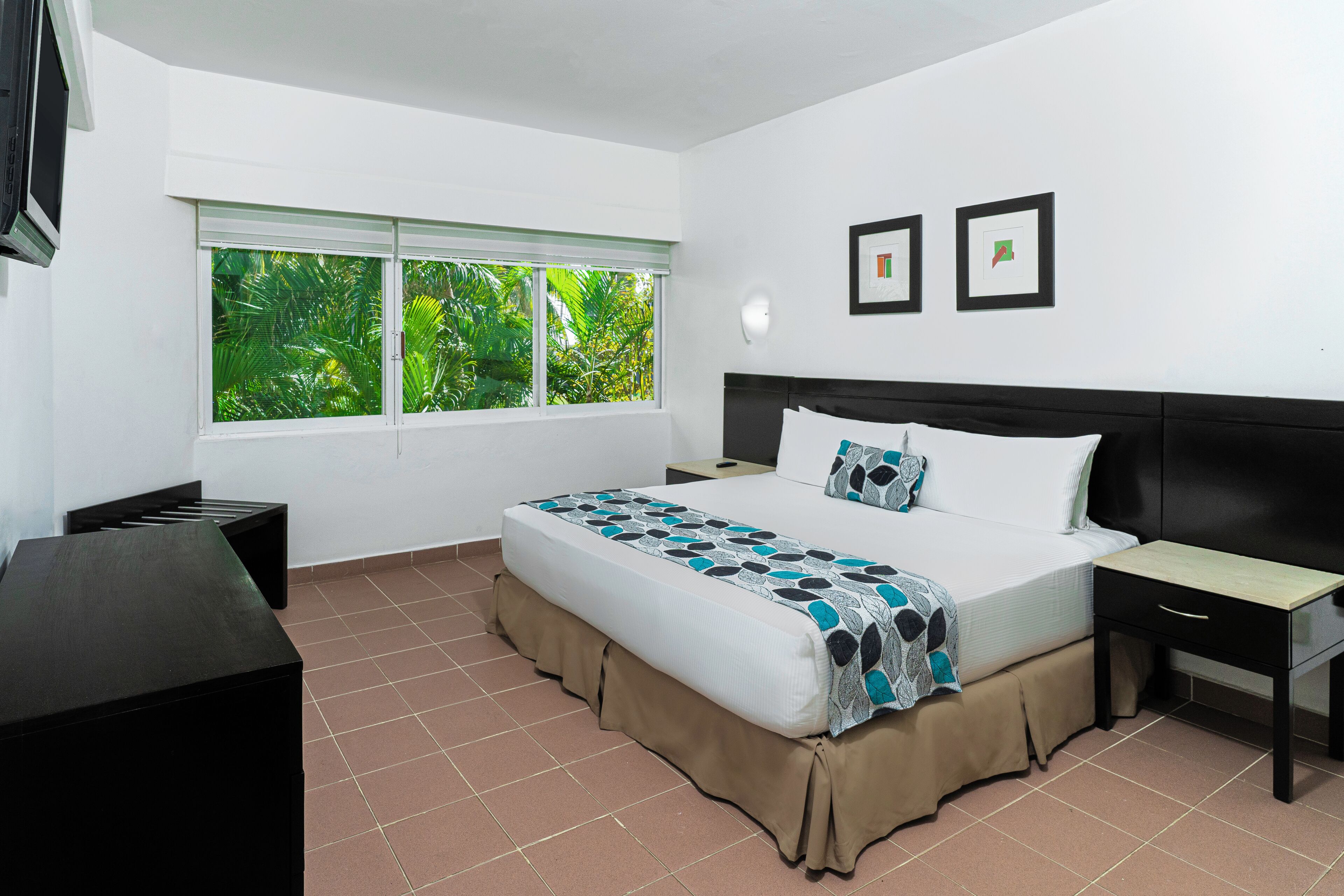 A hotel room with a bed, a desk, a chair, a window with a view of greenery, and two framed pictures on the wall.