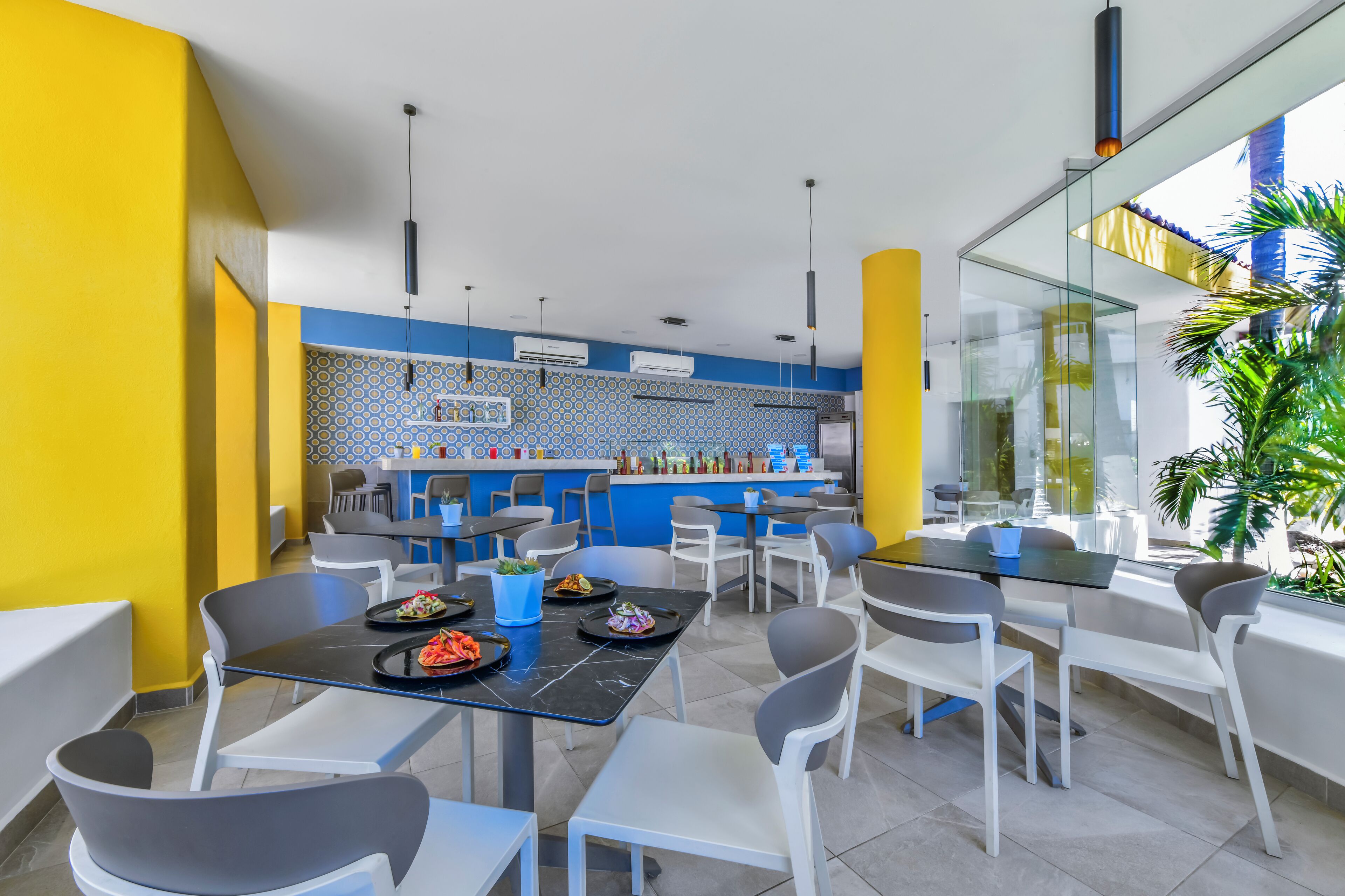 Un restaurante moderno con una paleta de colores brillante en amarillo y azul, que incluye una zona de bar, diversas opciones de asientos y vistas al exterior.
