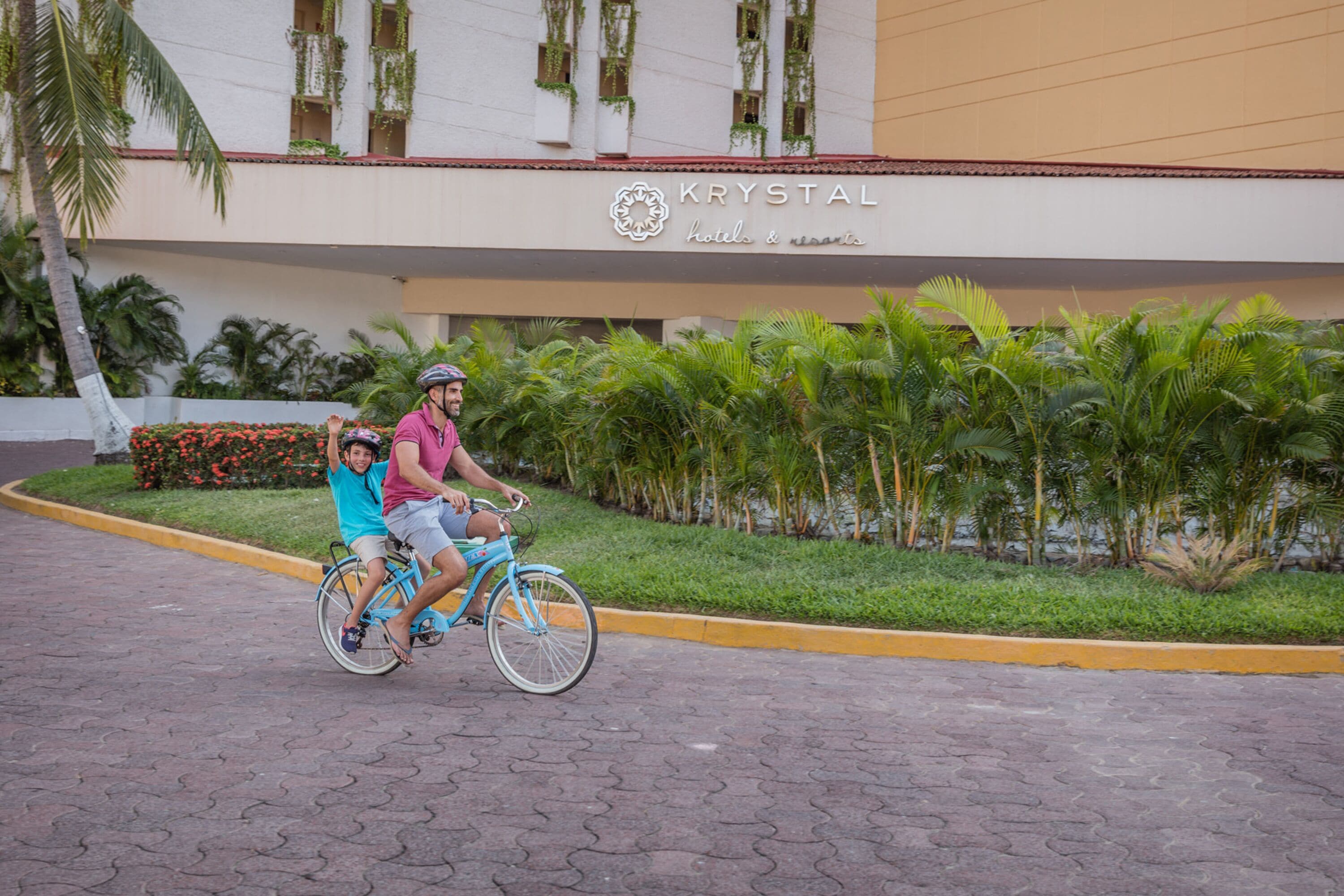 Two people riding a bicycle in front of a building with a sign that reads "KRYSTAL hotels & resorts."