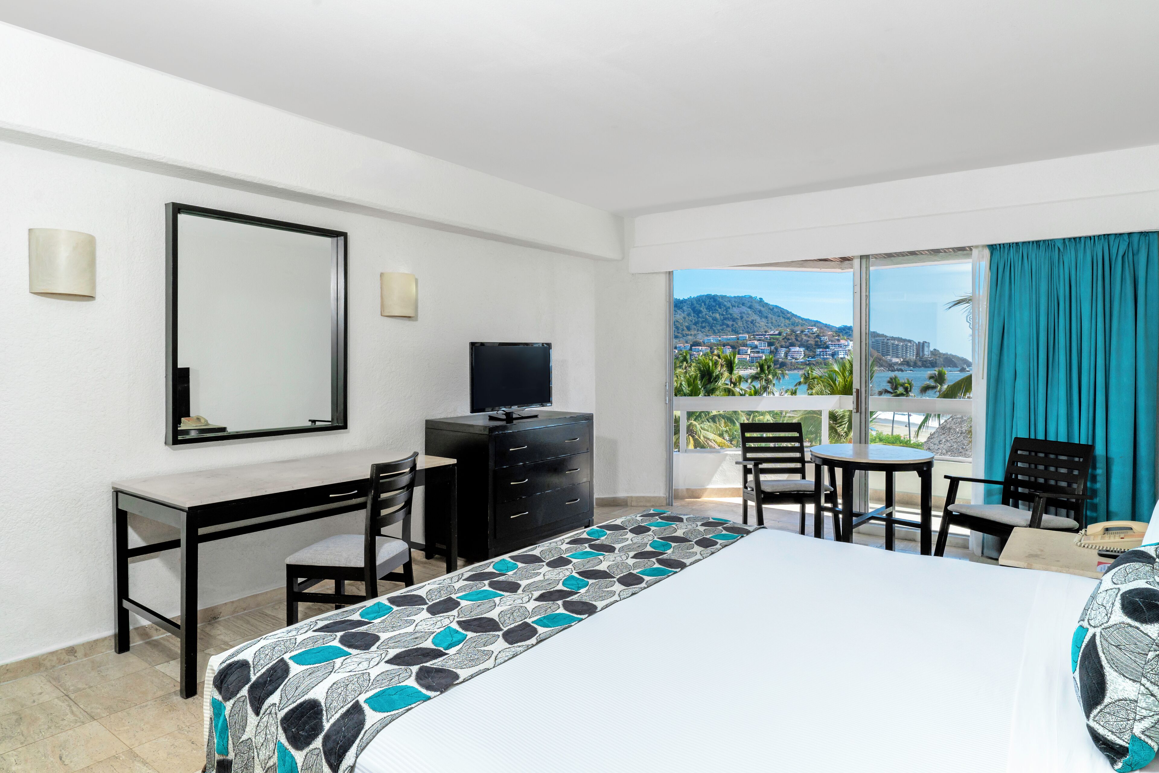 Ein Hotelzimmer mit Bett, Schreibtisch, Fernseher und einem Balkon mit Blick auf die Stadt und die Berge.