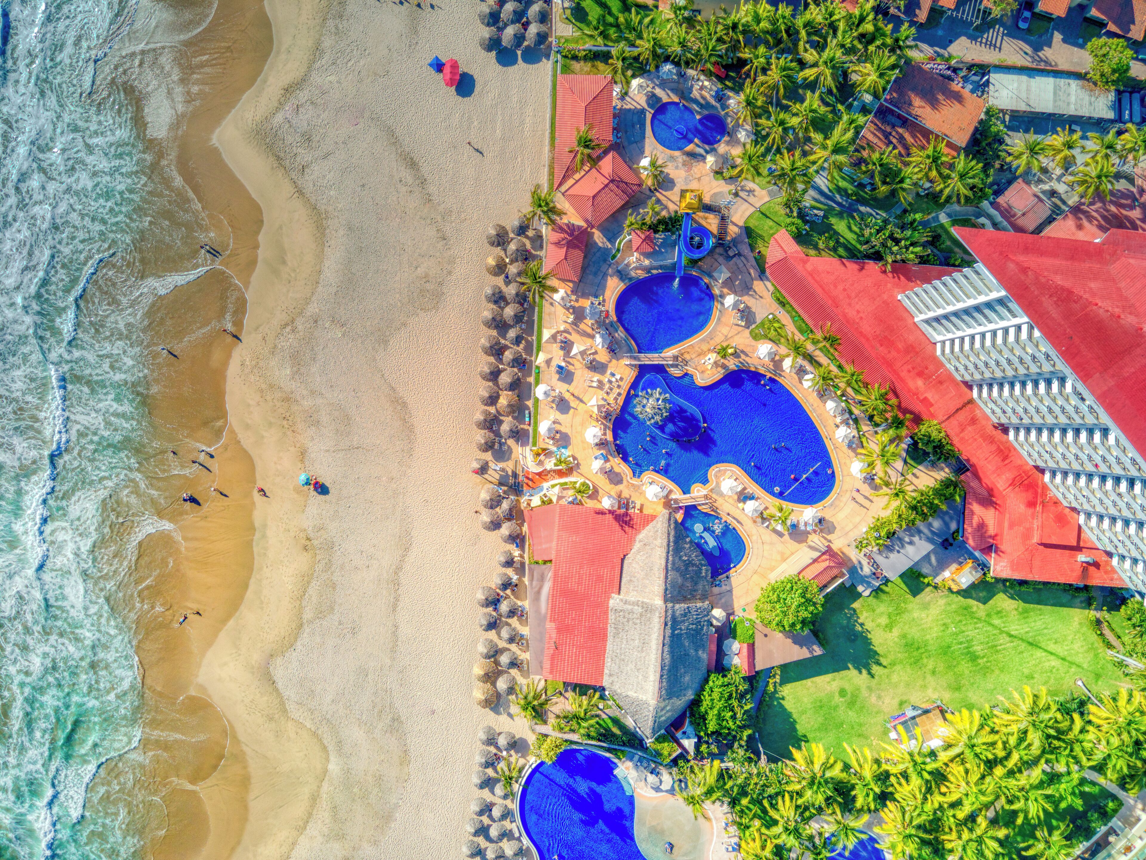 Vista aérea de un complejo turístico con varias piscinas, sombrillas de playa y palmeras.