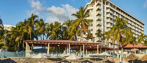A beachfront hotel with multiple floors, palm trees, and a beach area with sun loungers and umbrellas.