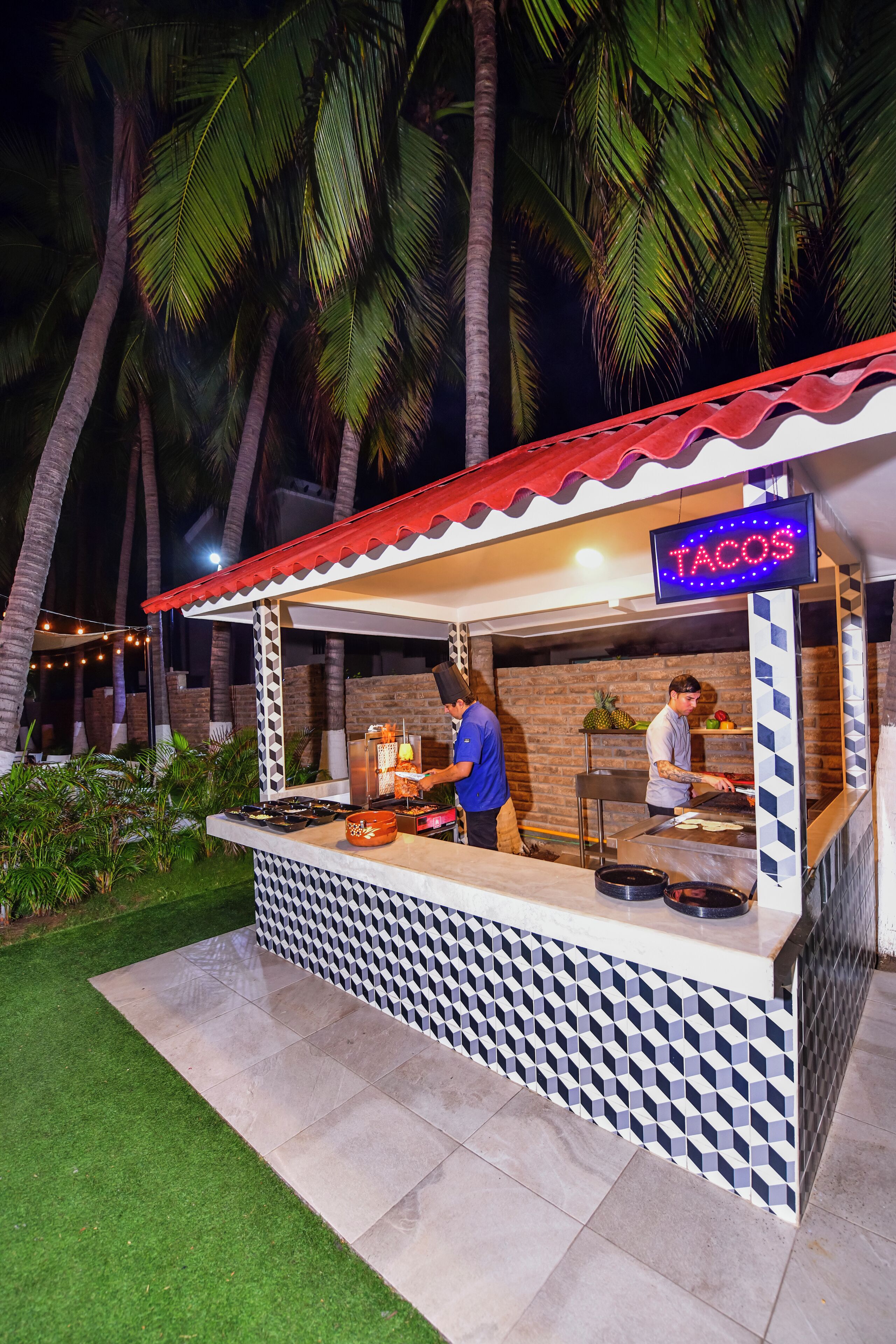 A taco stand with a neon sign, a tiled counter, and palm trees in the background.