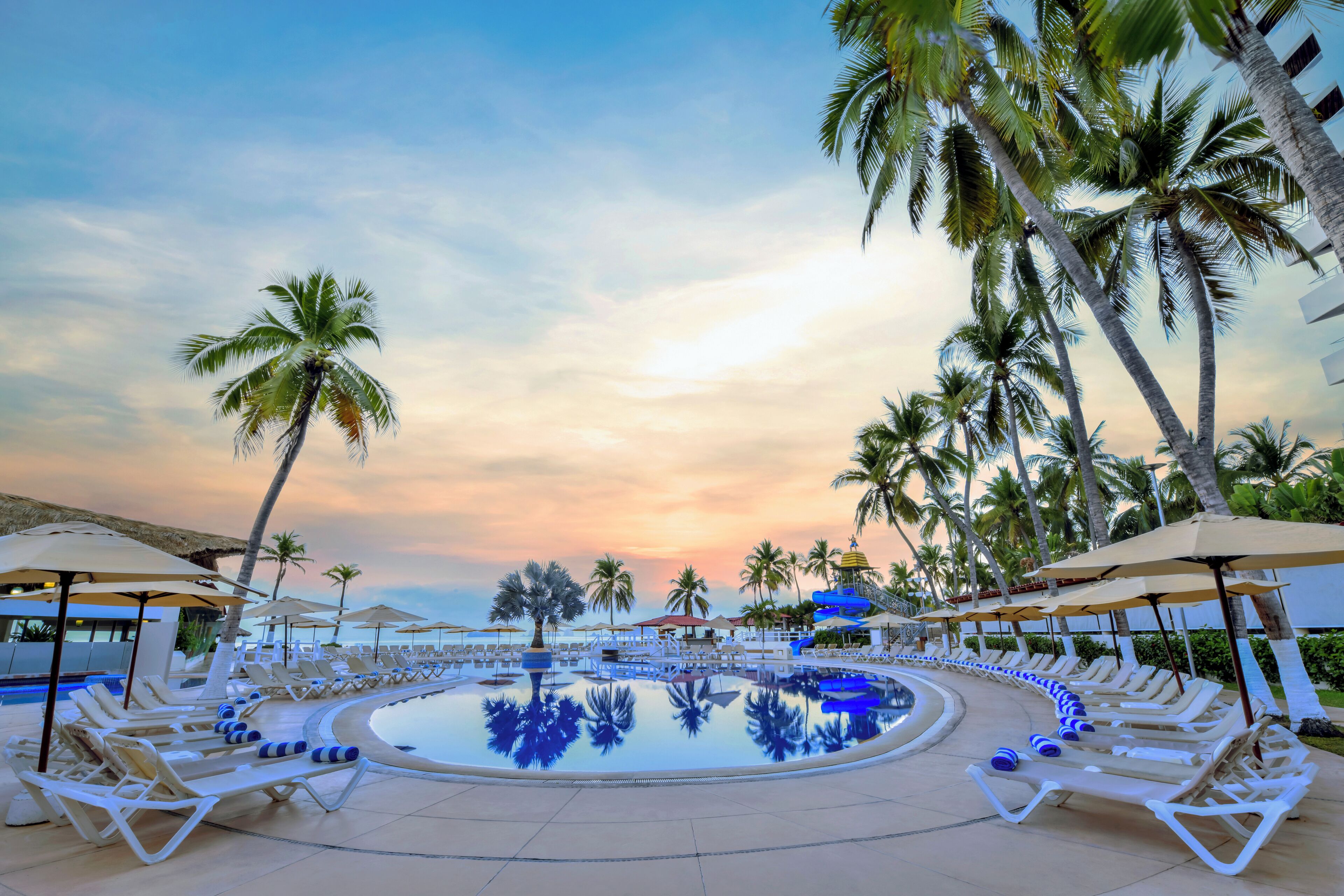 A resort pool area with lounge chairs and umbrellas, surrounded by palm trees, under a sunset sky.