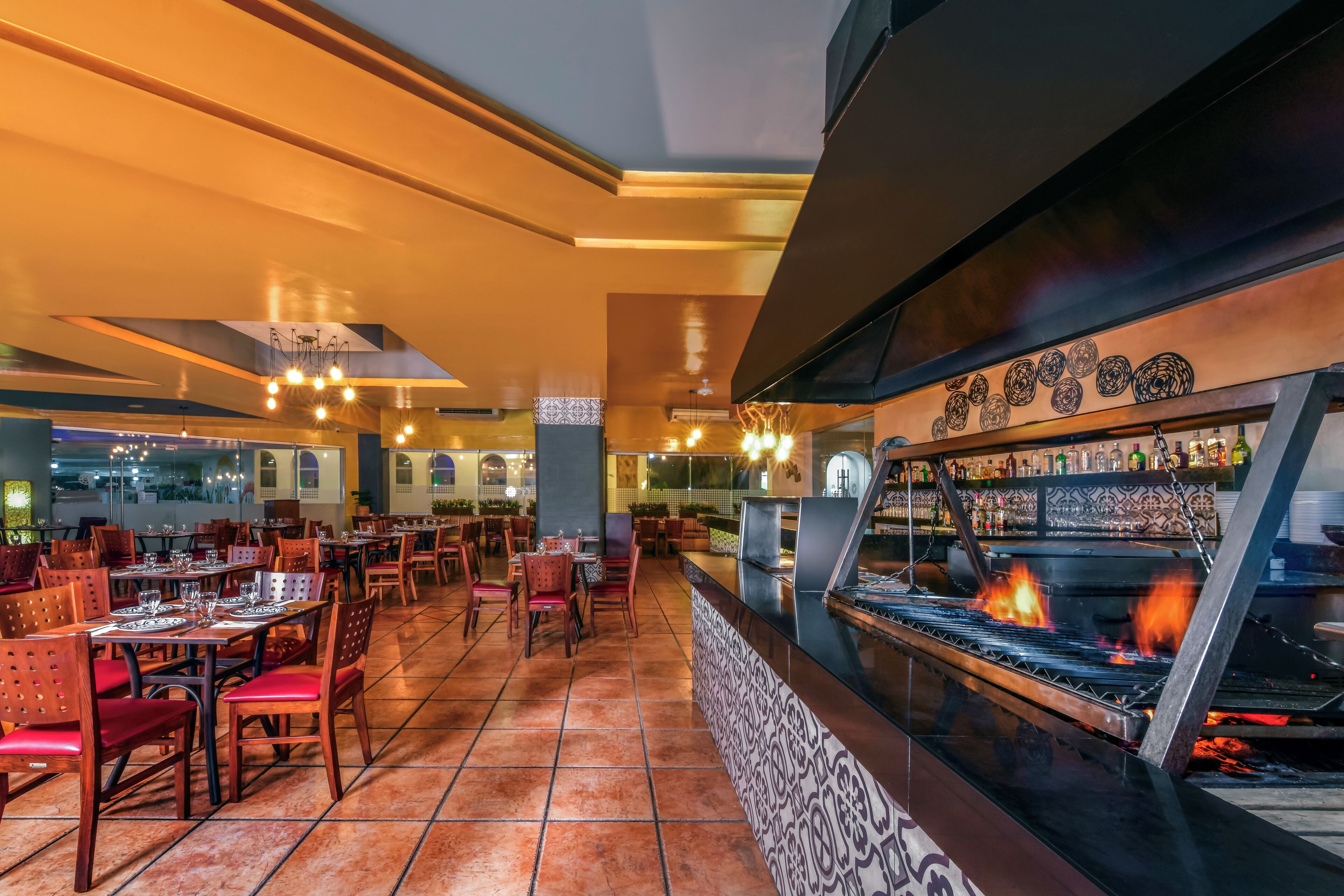 Restaurante con zona de bar y una parrilla. Mesas y sillas dispuestas para comer.