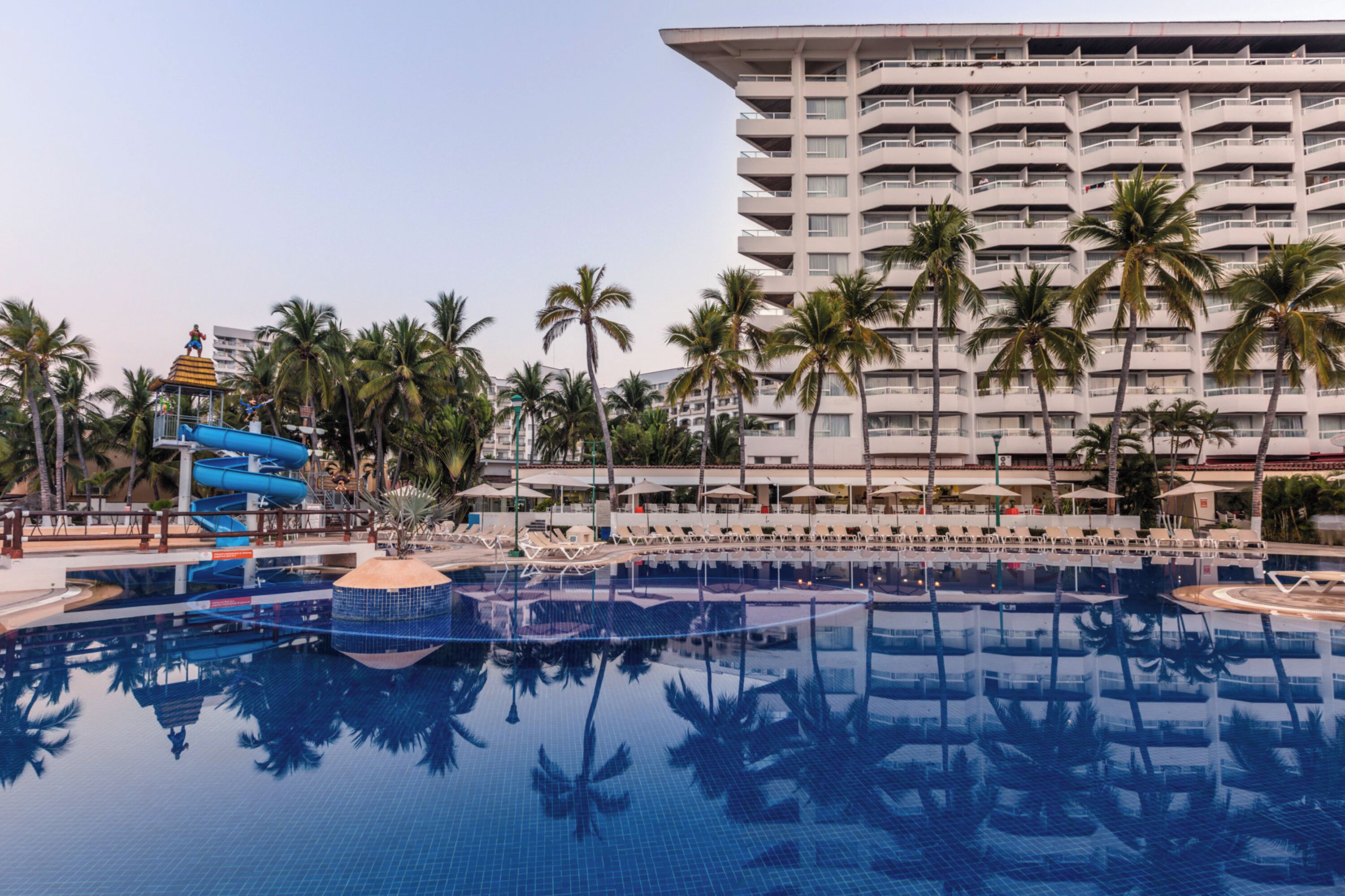 Zona de piscina hotelera con tobogán de agua, palmeras y un edificio grande al fondo.