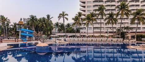 Zona de piscina hotelera con tobogán de agua, palmeras y un edificio grande al fondo.