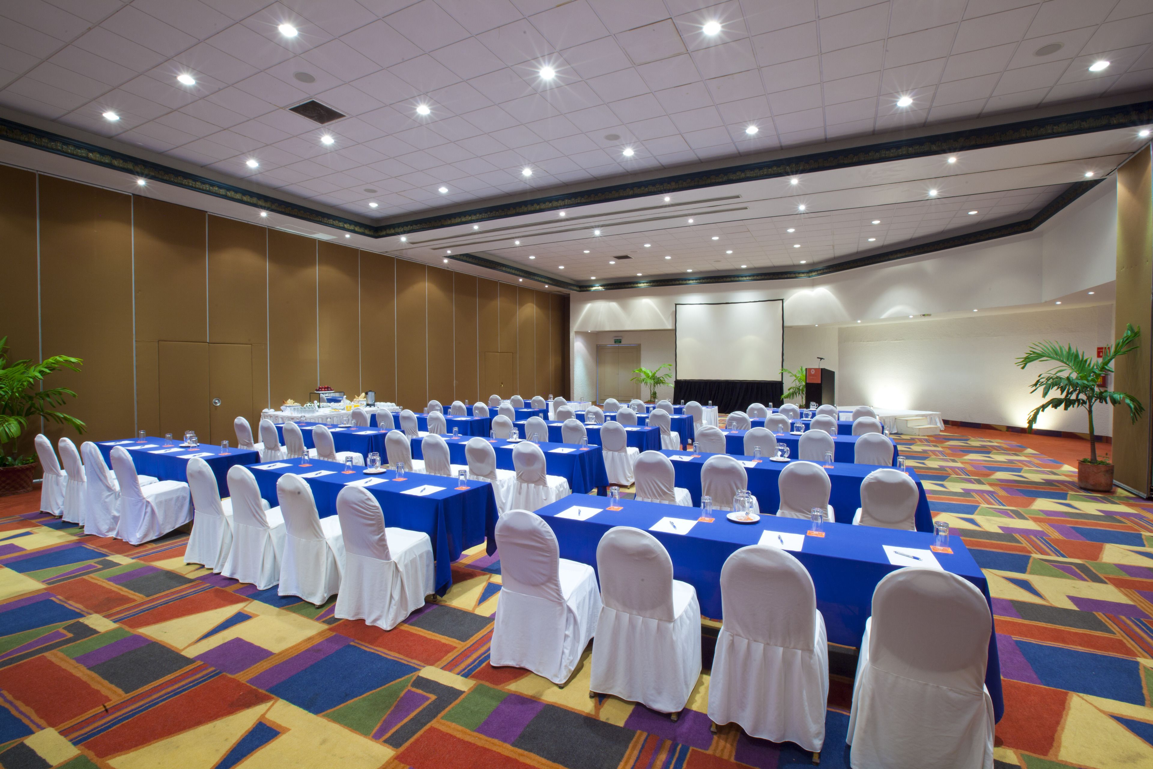 A conference room with multiple tables and chairs, arranged in rows, with a carpeted floor and a ceiling with recessed lighting.