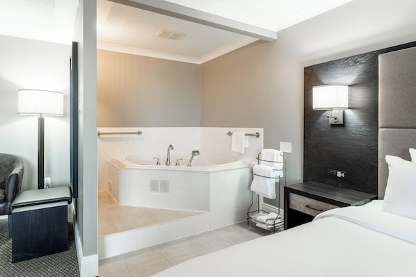 Une chambre d’hôtel moderne dotée d’un grand lit, d’une salle de bain séparée avec baignoire, d’un petit coin salon et d’une table de chevet avec une lampe.