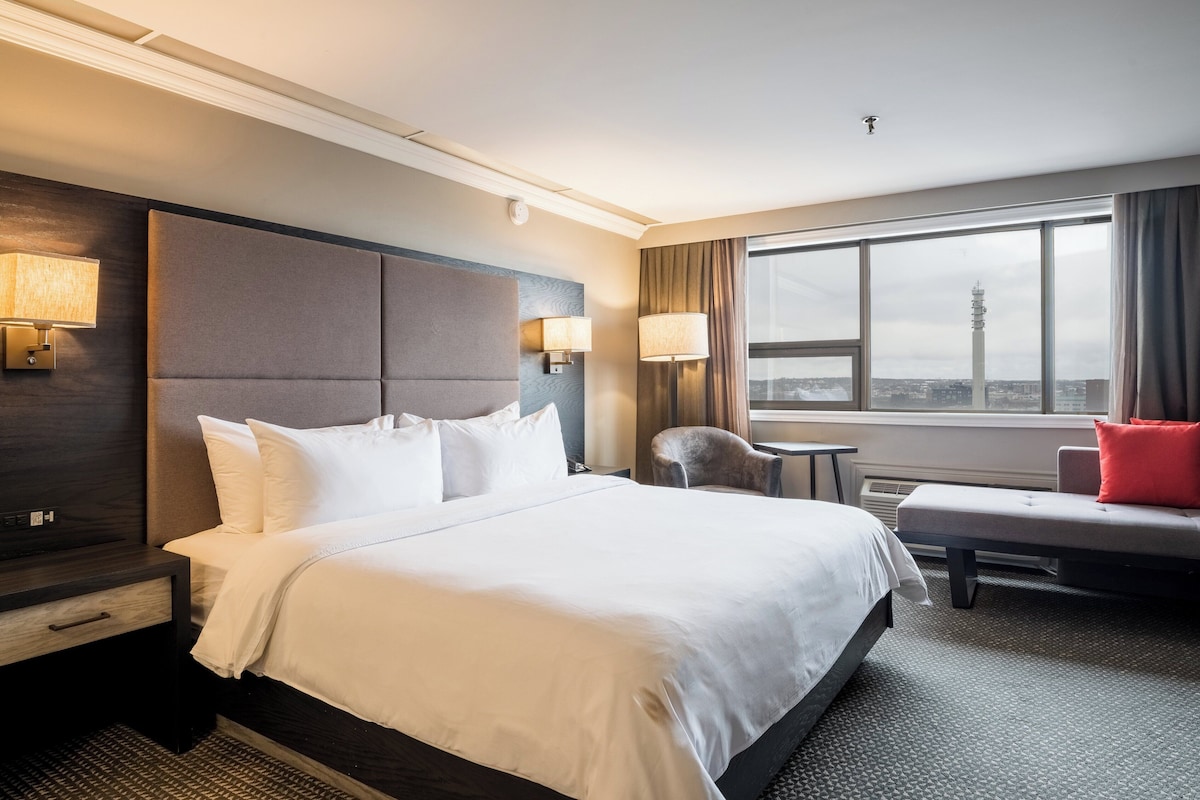 A hotel room with a large bed, a chair, a small table, and a view of a cityscape through a large window.