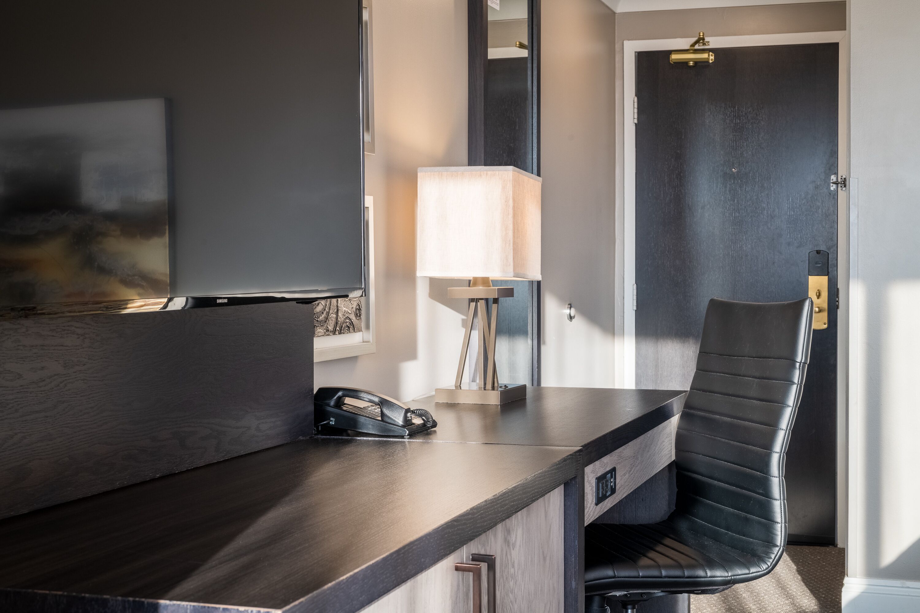 Une chambre d’hôtel moderne avec un bureau en bois foncé, un fauteuil en cuir noir, une lampe et un téléphone.