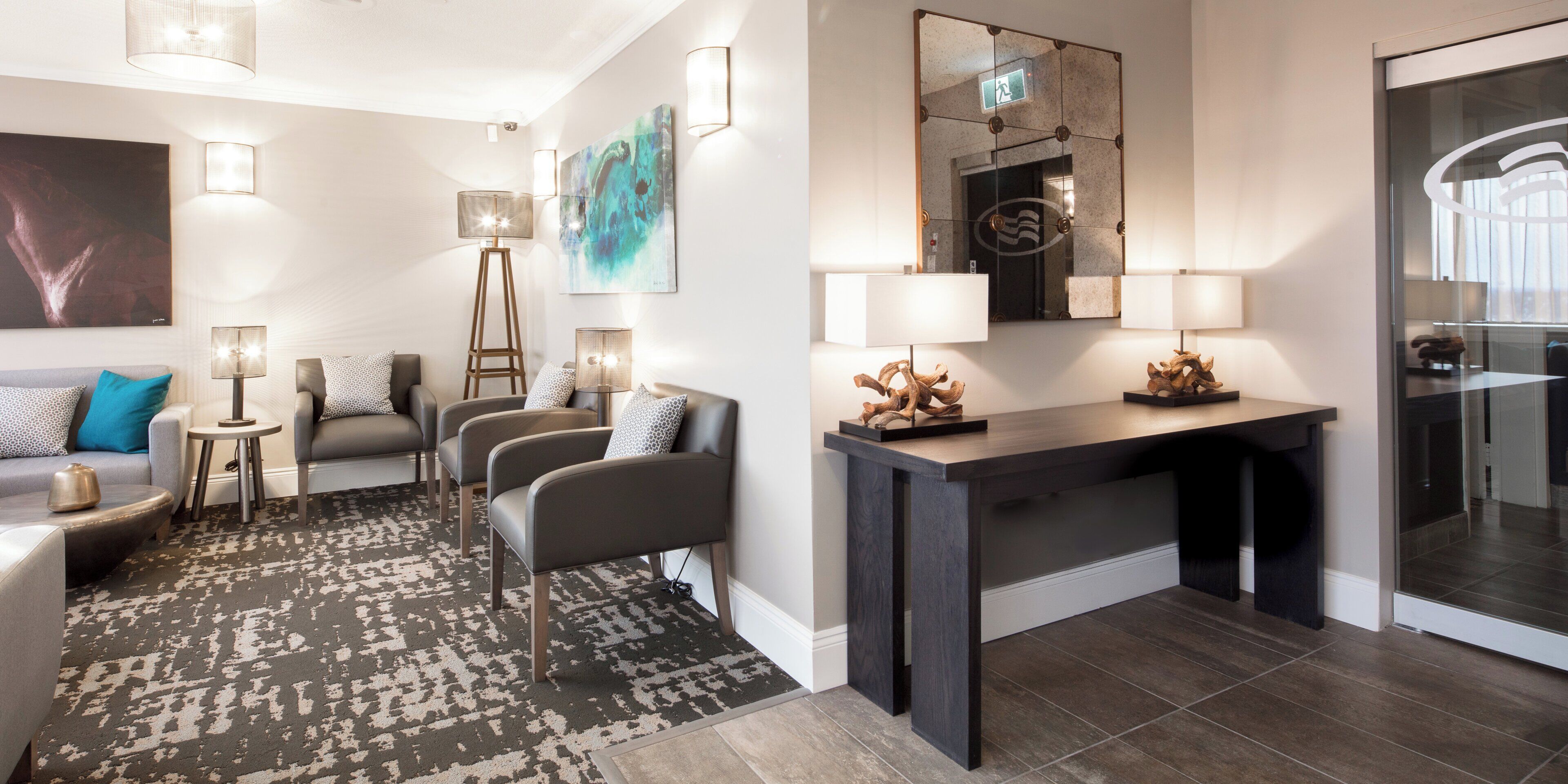 A modern hotel room with a sofa, armchairs, a coffee table, a desk, and a mirror.